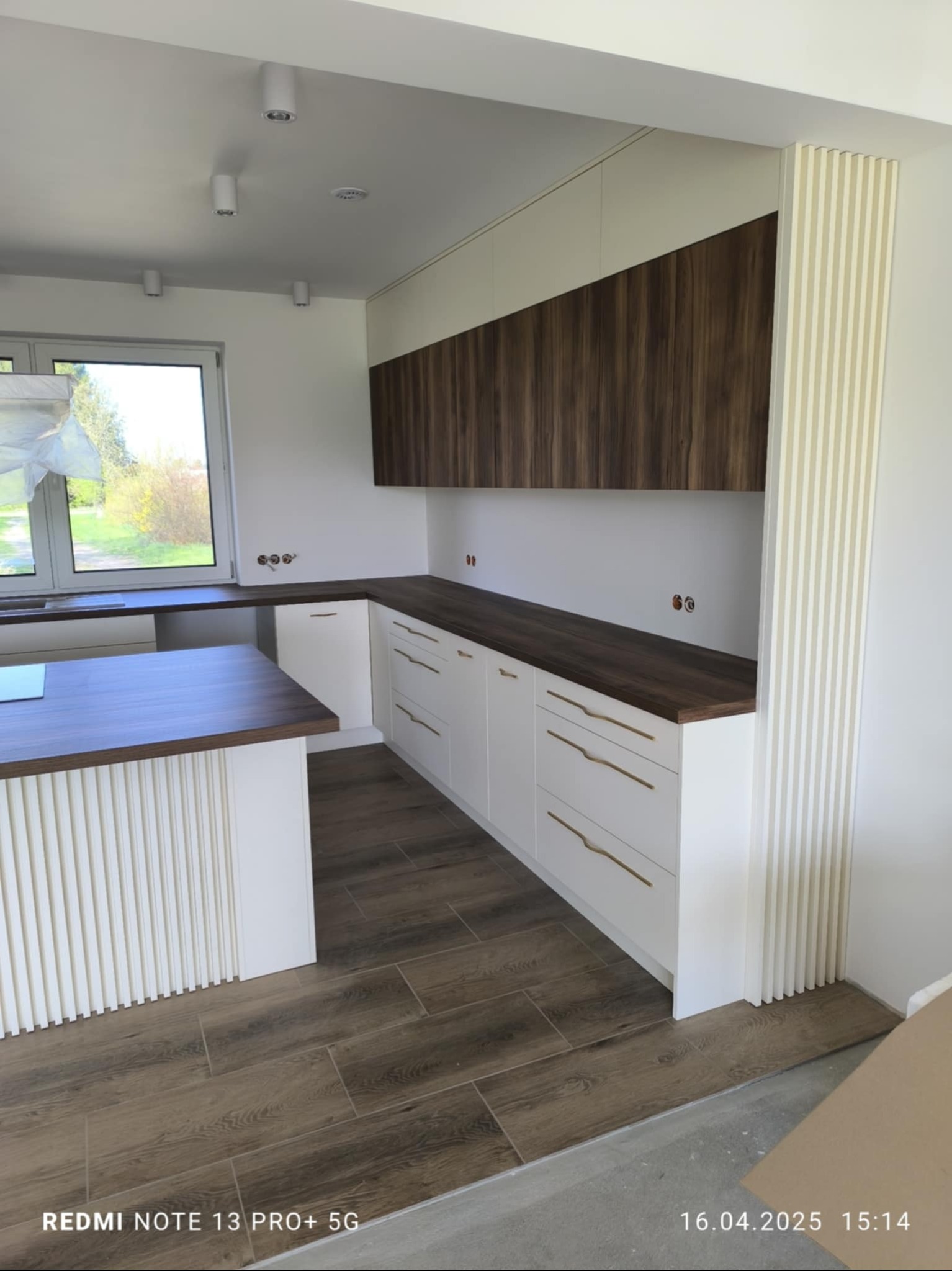 Nowoczesna kuchnia w bieli i drewnie: dolne szafki z złotymi uchwytami, drewniany blat i górne szafki. Na podłodze panele imitujące drewno. Widok na wyspę kuchenną.