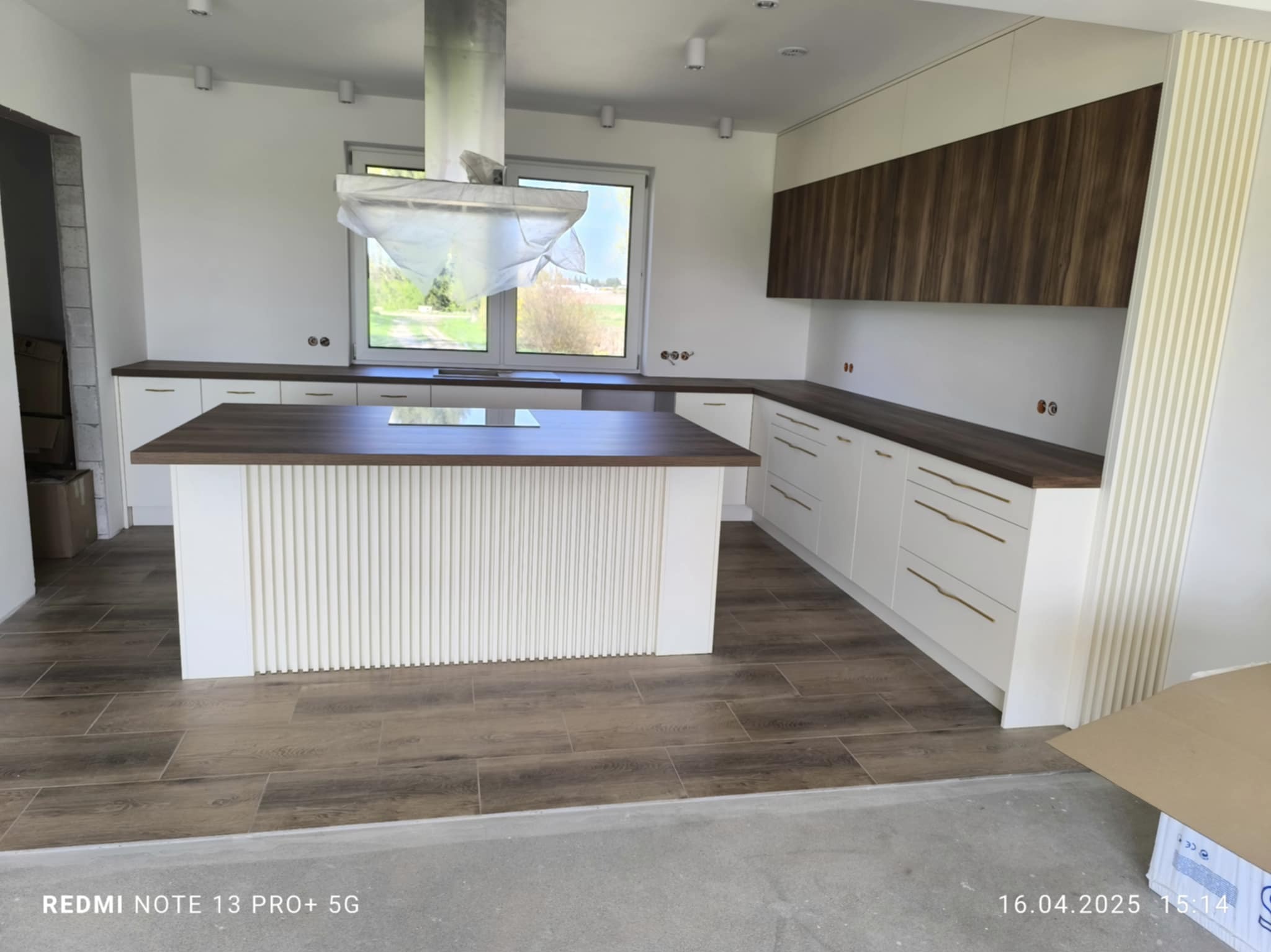 Nowoczesna kuchnia z wyspą, białe szafki z drewnianymi blatami, okap w folii ochronnej. Podłoga w panelach imitujących drewno. Minimalistyczny design.