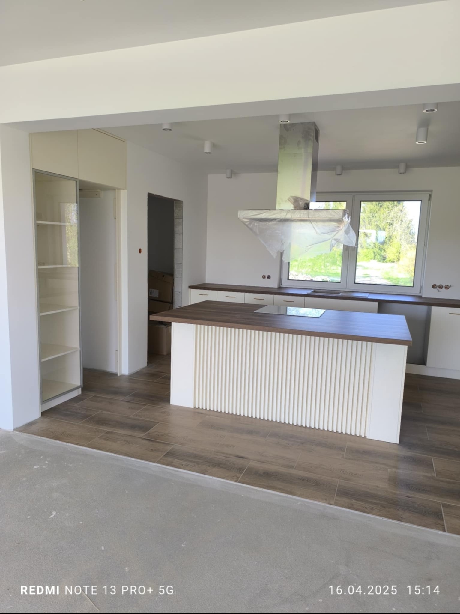 Nowoczesna, jasna kuchnia z wyspą i okapem w folii ochronnej. Podłoga wyłożona panelami imitującymi drewno. Widoczna zabudowa kuchenna z frontami bez uchwytów.