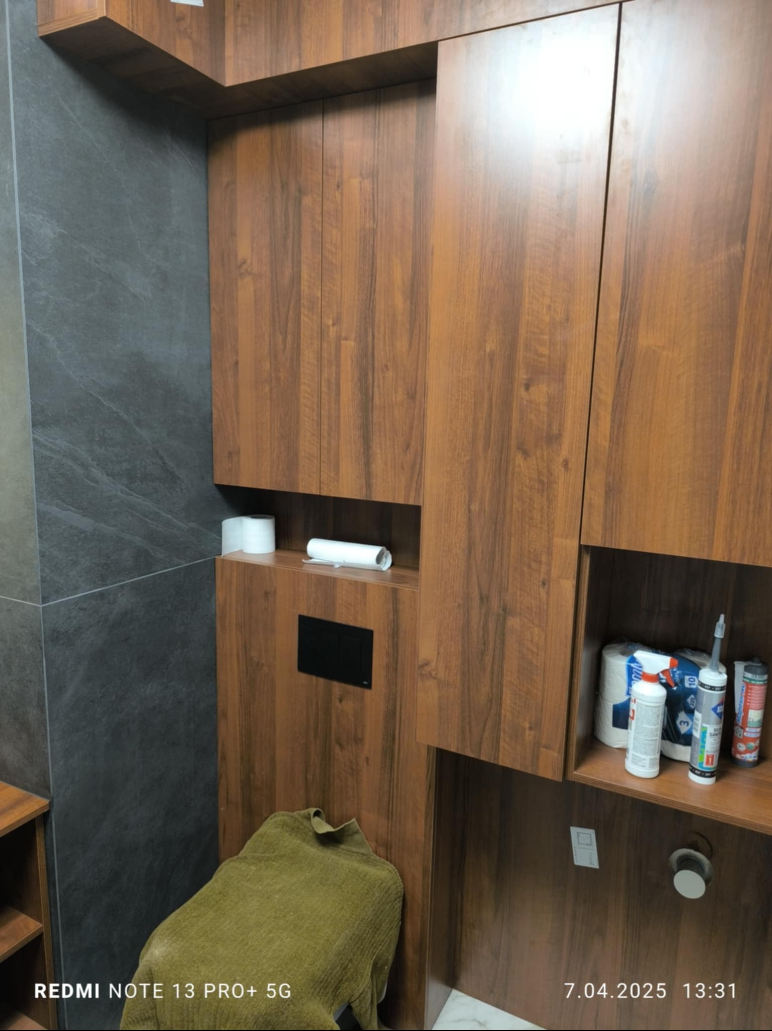 Drewniane szafki łazienkowe z ukrytą spłuczką i półkami na akcesoria toaletowe, na tle szarej ściany z płytek. Widoczny fragment toalety z ręcznikiem.