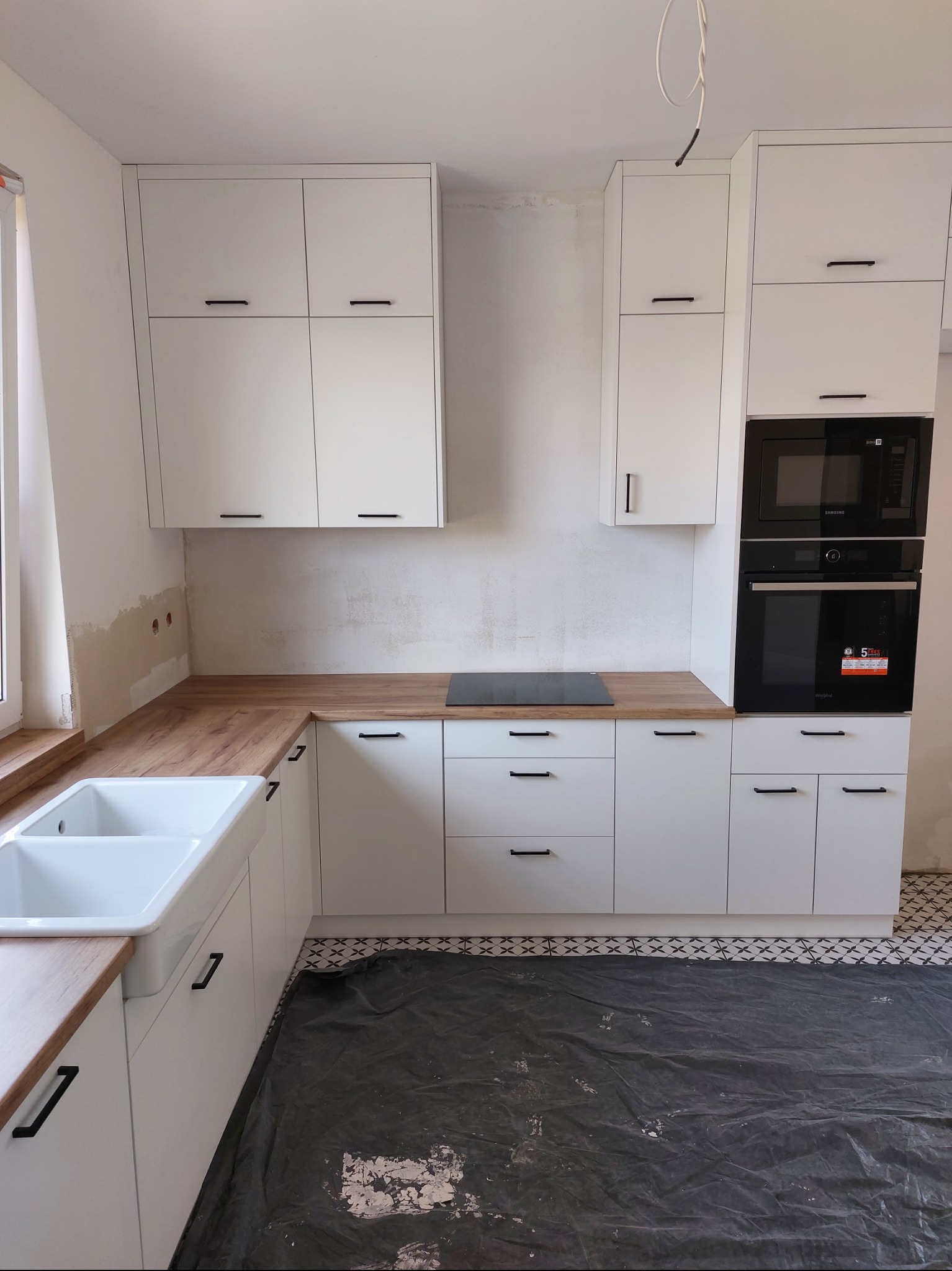 Biała kuchnia z drewnianym blatem i czarnymi uchwytami, piekarnik w zabudowie. Podłoga zabezpieczona folią malarską, ściany częściowo nie wykończone.