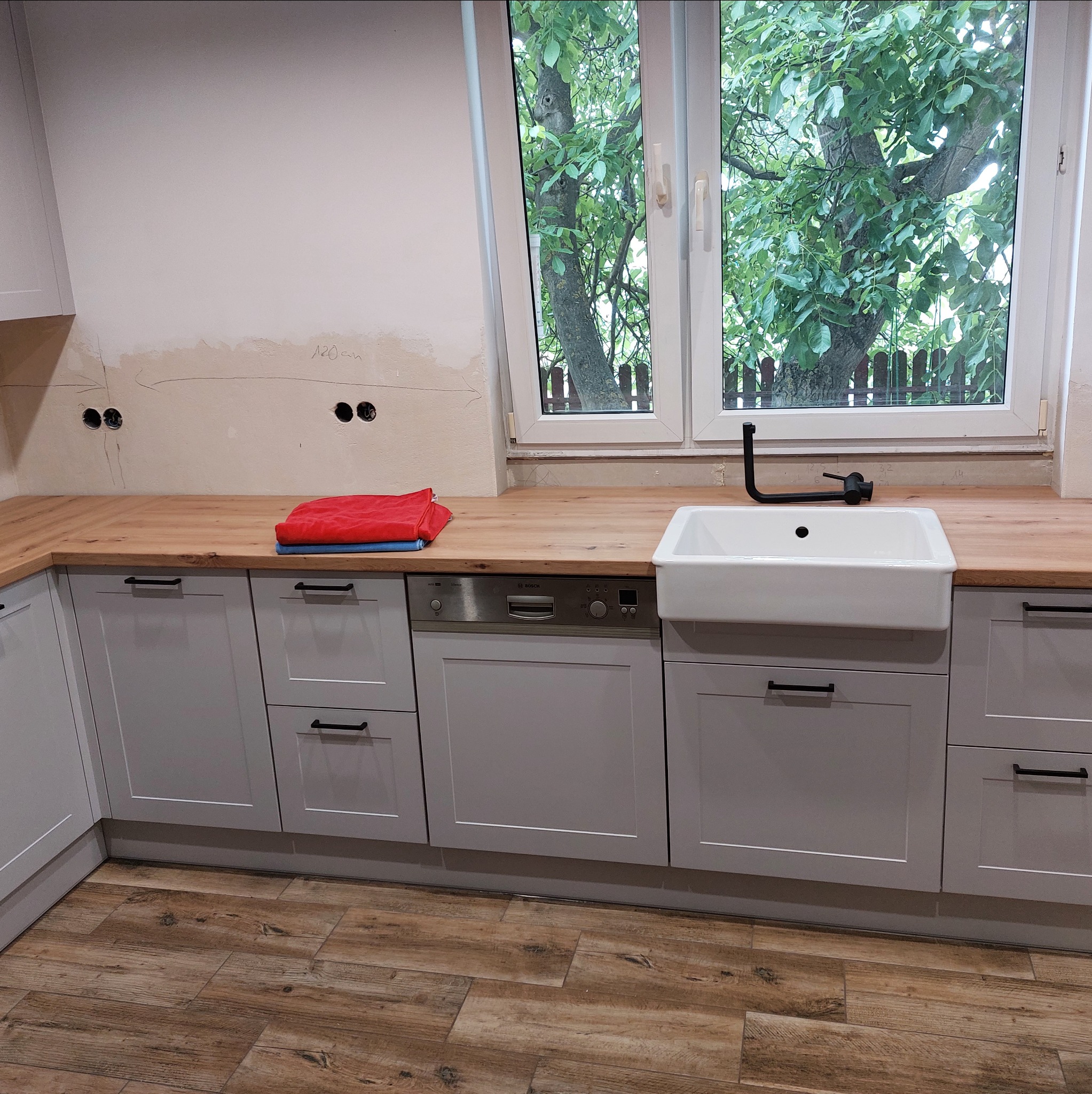 Szare szafki kuchenne z drewnianym blatem i białym zlewem farmerskim pod oknem z widokiem na drzewo. Częściowo wykończona ściana z widocznymi puszkami elektrycznymi.