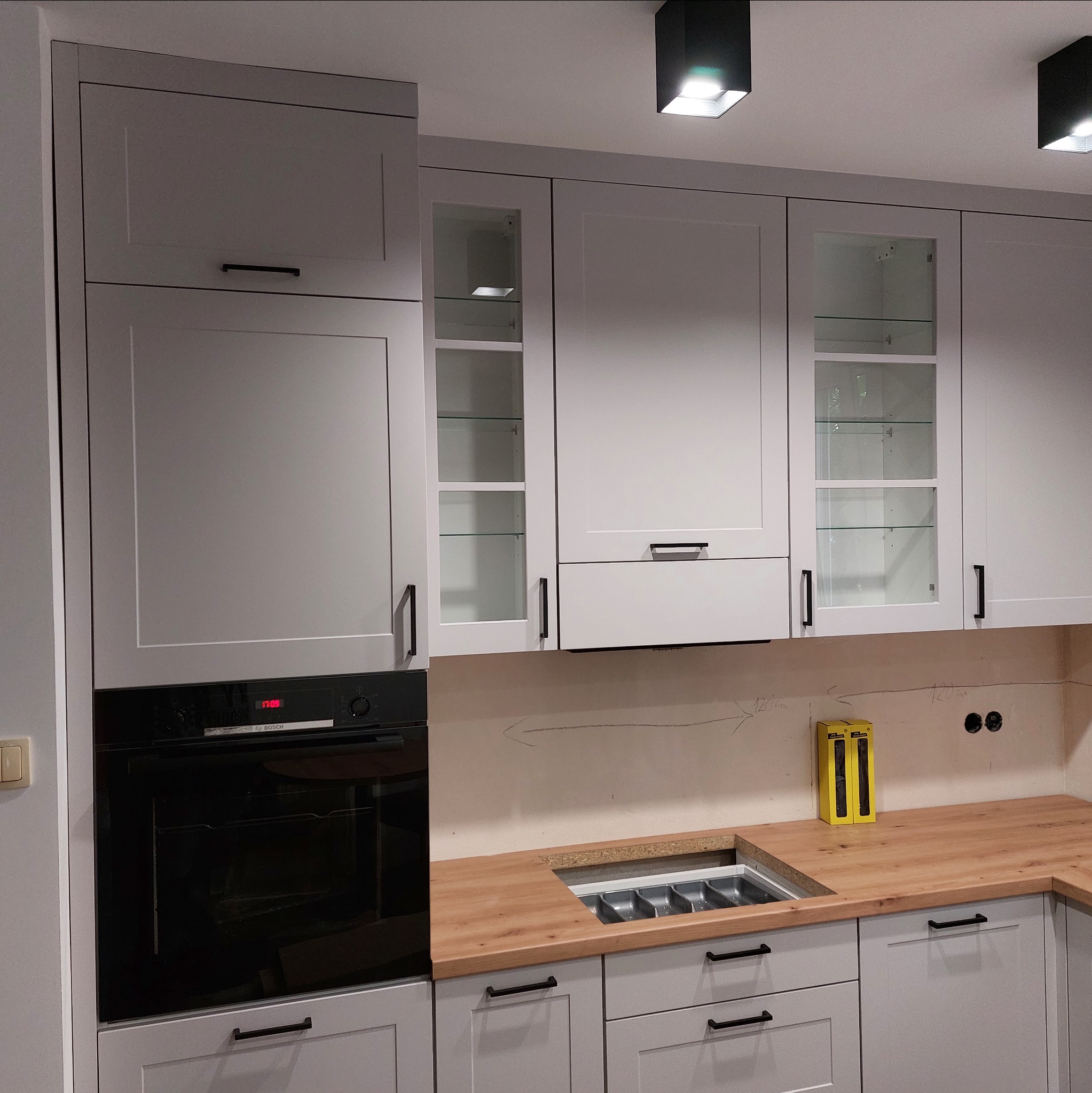 Szare szafki kuchenne z czarnymi uchwytami i szklanymi witrynami, zabudowany piekarnik, drewniany blat z wkładem na sztućce, czarne lampy sufitowe.