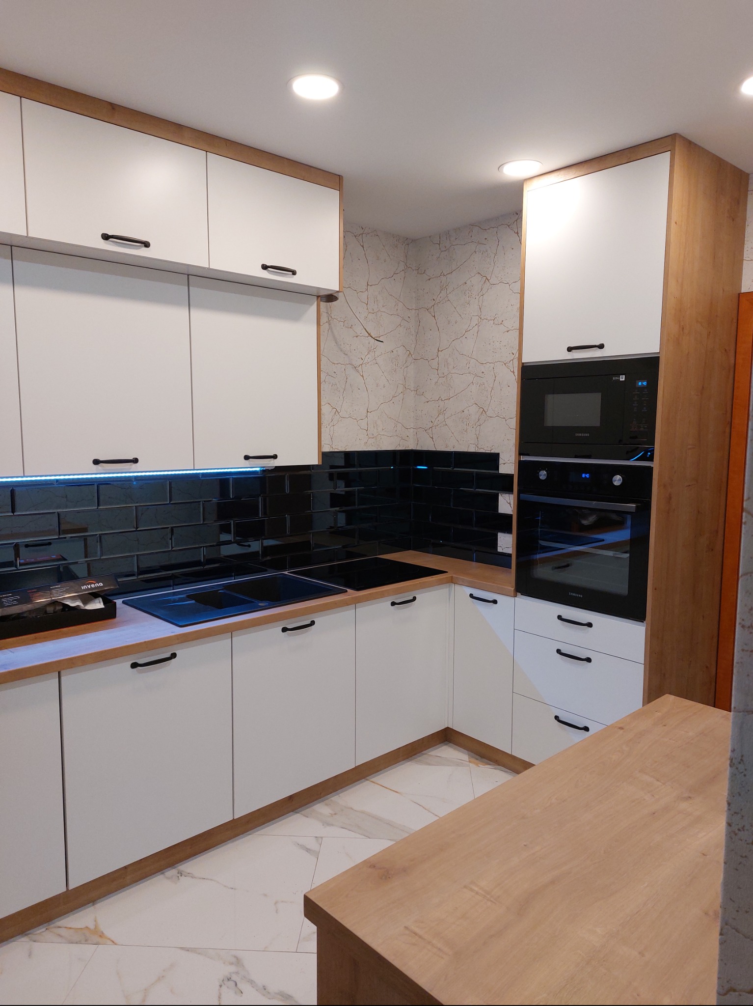 Nowoczesna kuchnia z białymi frontami i drewnianymi akcentami. Czarne płytki nad blatem. Oświetlenie LED. Widoczne sprzęty AGD Samsung.