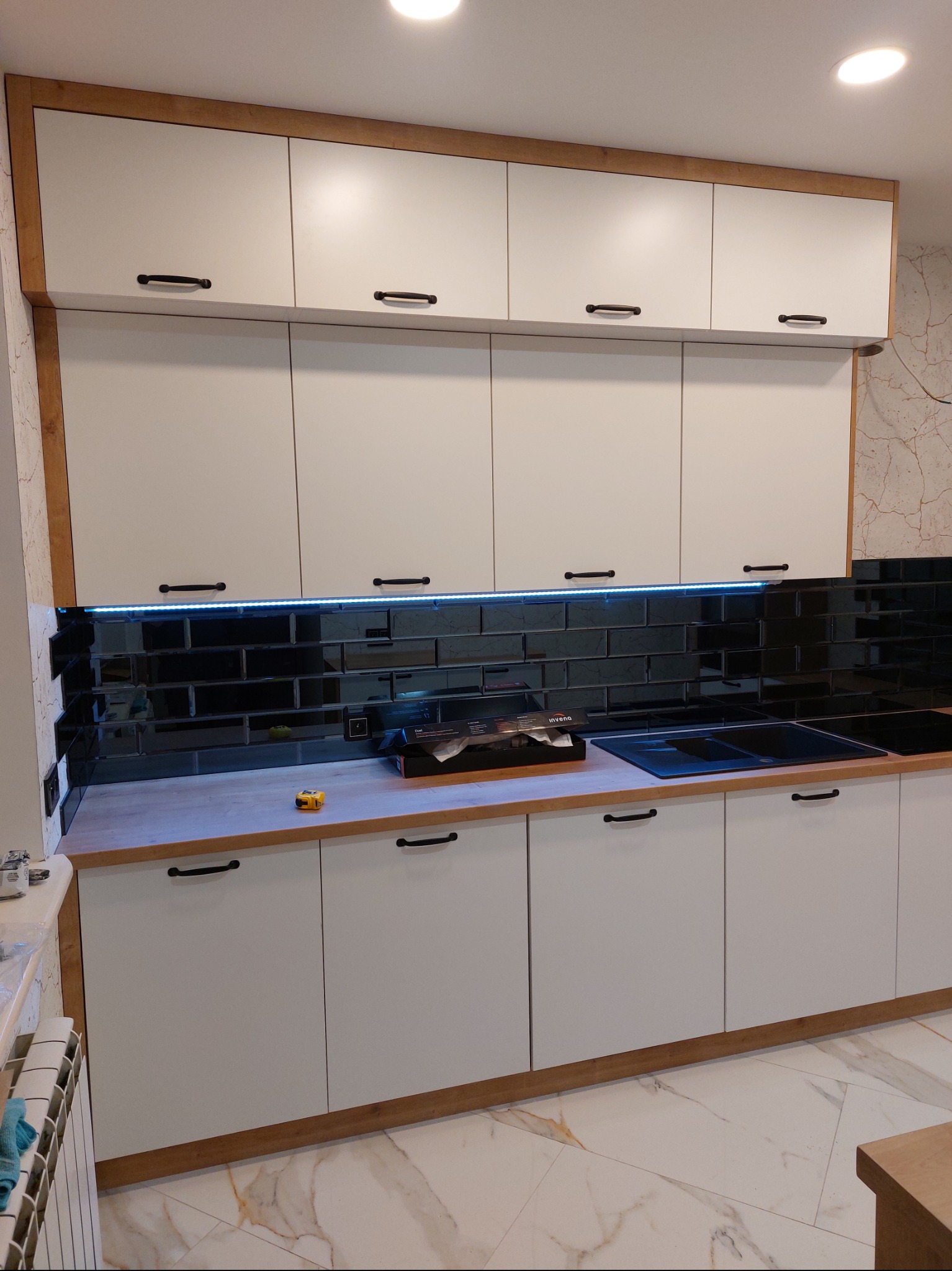 Biała zabudowa kuchenna z czarnymi uchwytami i lustrzanymi płytkami nad blatem, podświetlona listwą LED. Uwagę zwraca wykończenie w kolorze drewna.