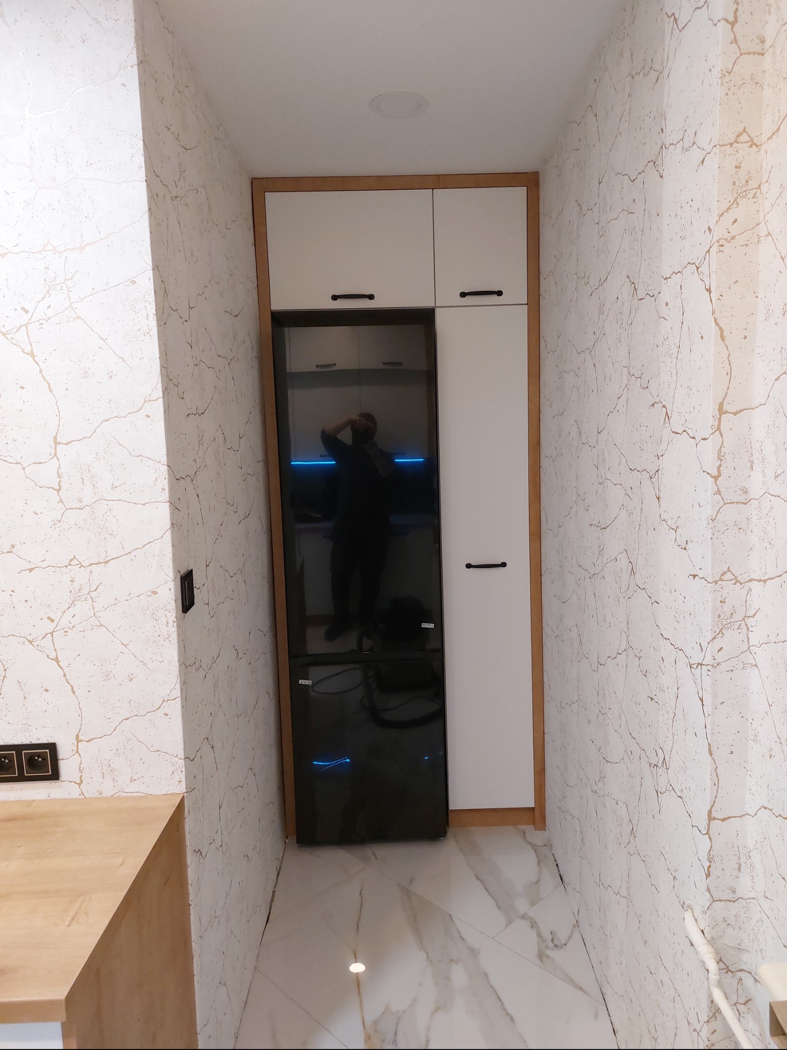 Nowoczesna zabudowa kuchenna z białymi szafkami i czarną lodówką, widok na wąskie przejście z marmurową podłogą i ścianami w beżowe żyłki, odbicie osoby w lodówce.