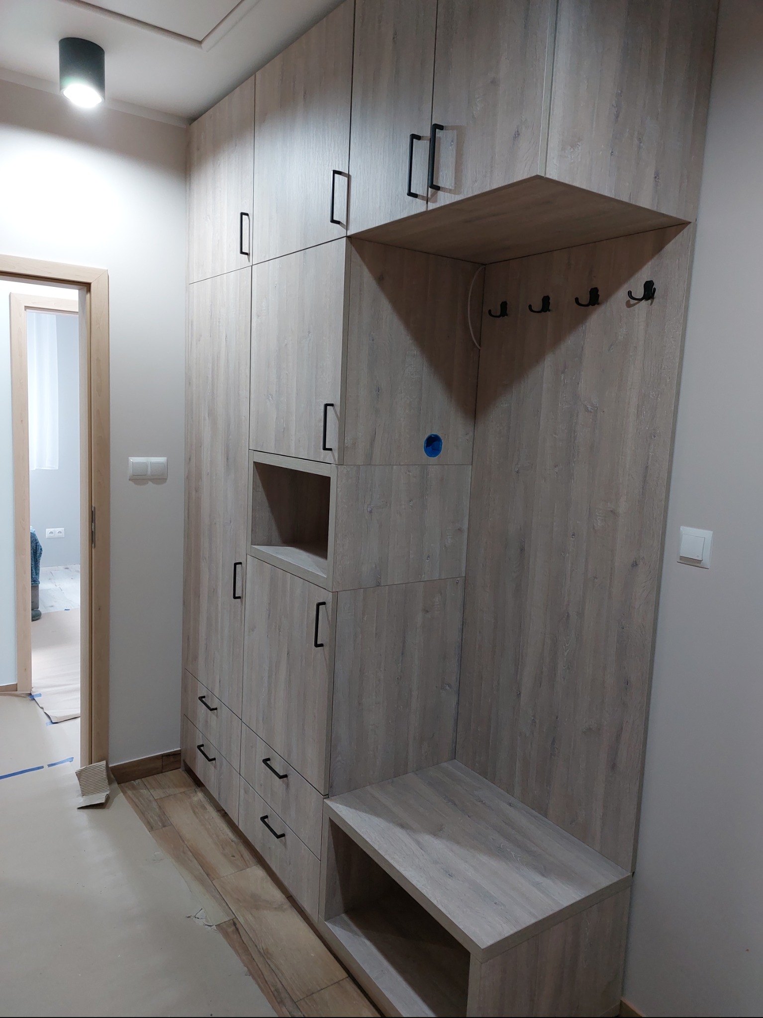 Szafa wnękowa z siedziskiem i wieszakami, fronty w odcieniach drewna, czarne uchwyty, nowoczesny design, oświetlenie sufitowe.