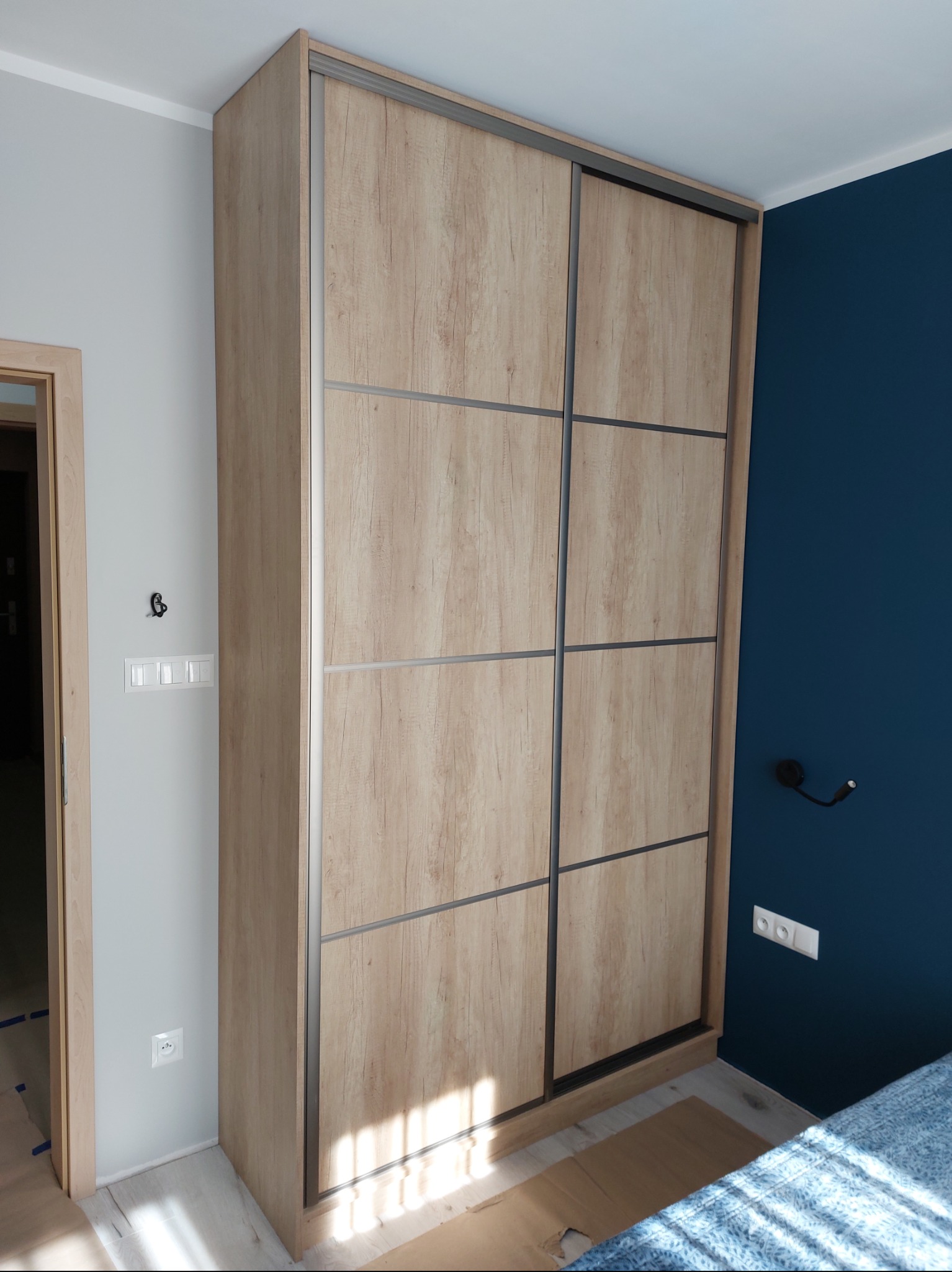 Jasna szafa wnękowa z przesuwnymi drzwiami o drewnianej fakturze, z ciemnymi obramowaniami, na tle ściany w kolorze głębokiego granatu i jasnoszarego.
