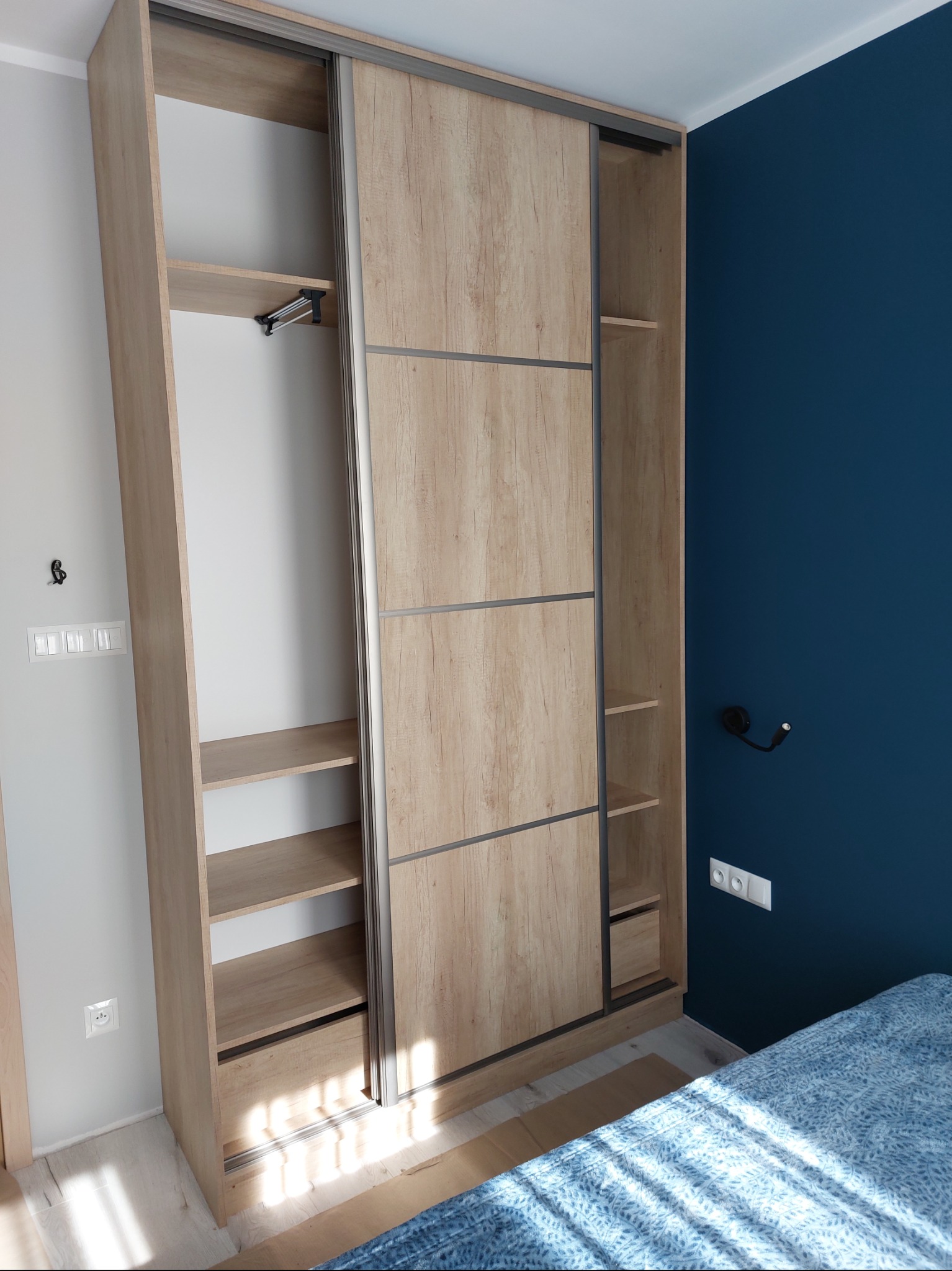 Szafa wnękowa z przesuwnymi drzwiami o strukturze drewna, z widocznym wnętrzem i półkami, na tle ściany w kolorze głębokiego granatu.