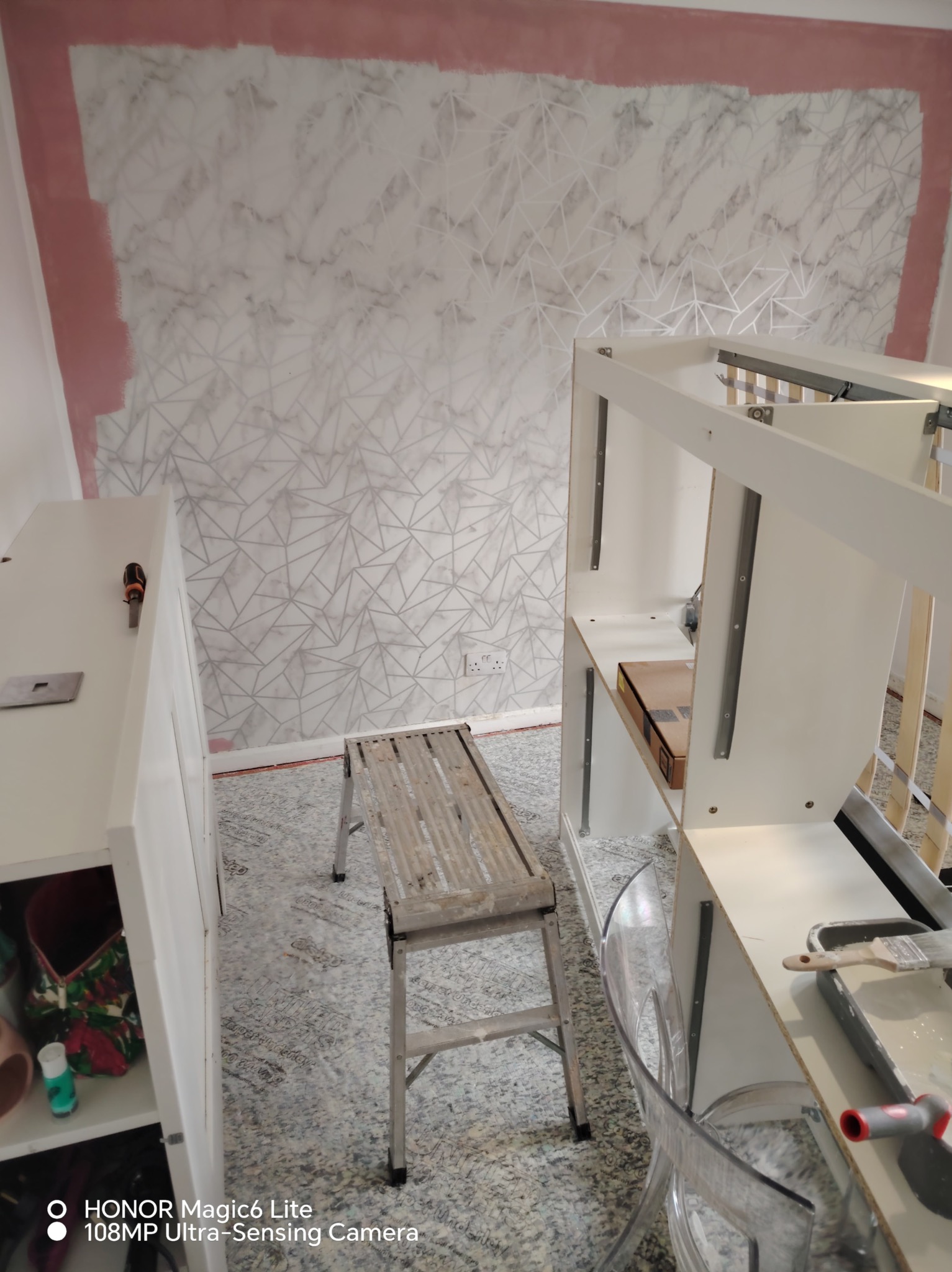 Pokój w trakcie malowania: ściana z geometrycznym wzorem, pomalowane obrzeże, drabina, rozłożone meble do montażu, kuweta z farbą i pędzel.