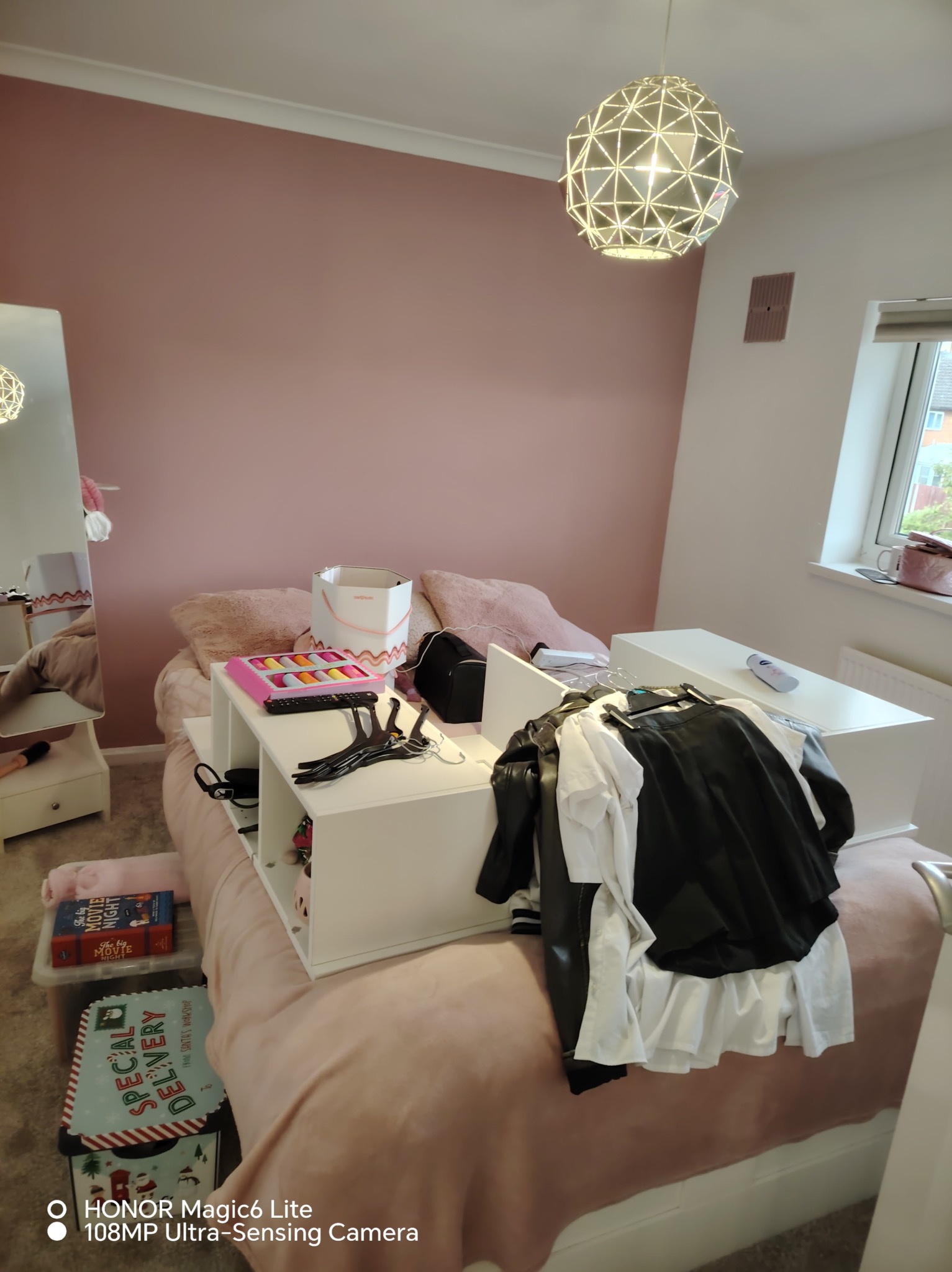 Różowa sypialnia przed remontem: białe meble z ubraniami na łóżku, pudrowa ściana, lampa sufitowa. Widoczny bałagan przed wykończeniem wnętrza.