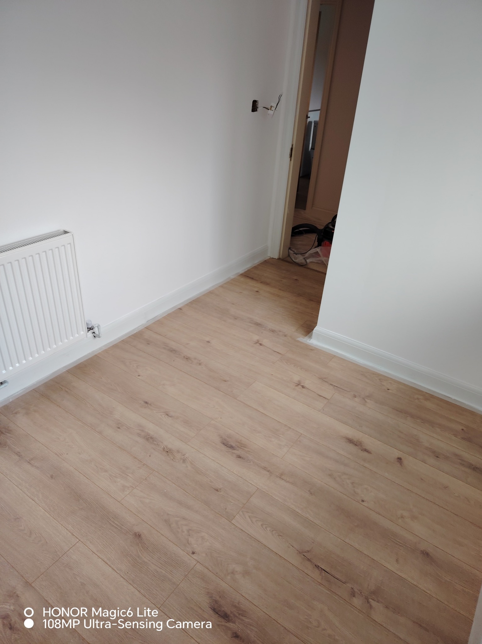 Jasne panele podłogowe w pokoju z białymi ścianami i grzejnikiem. Widoczne listwy przypodłogowe i otwarte drzwi do kolejnego pomieszczenia.