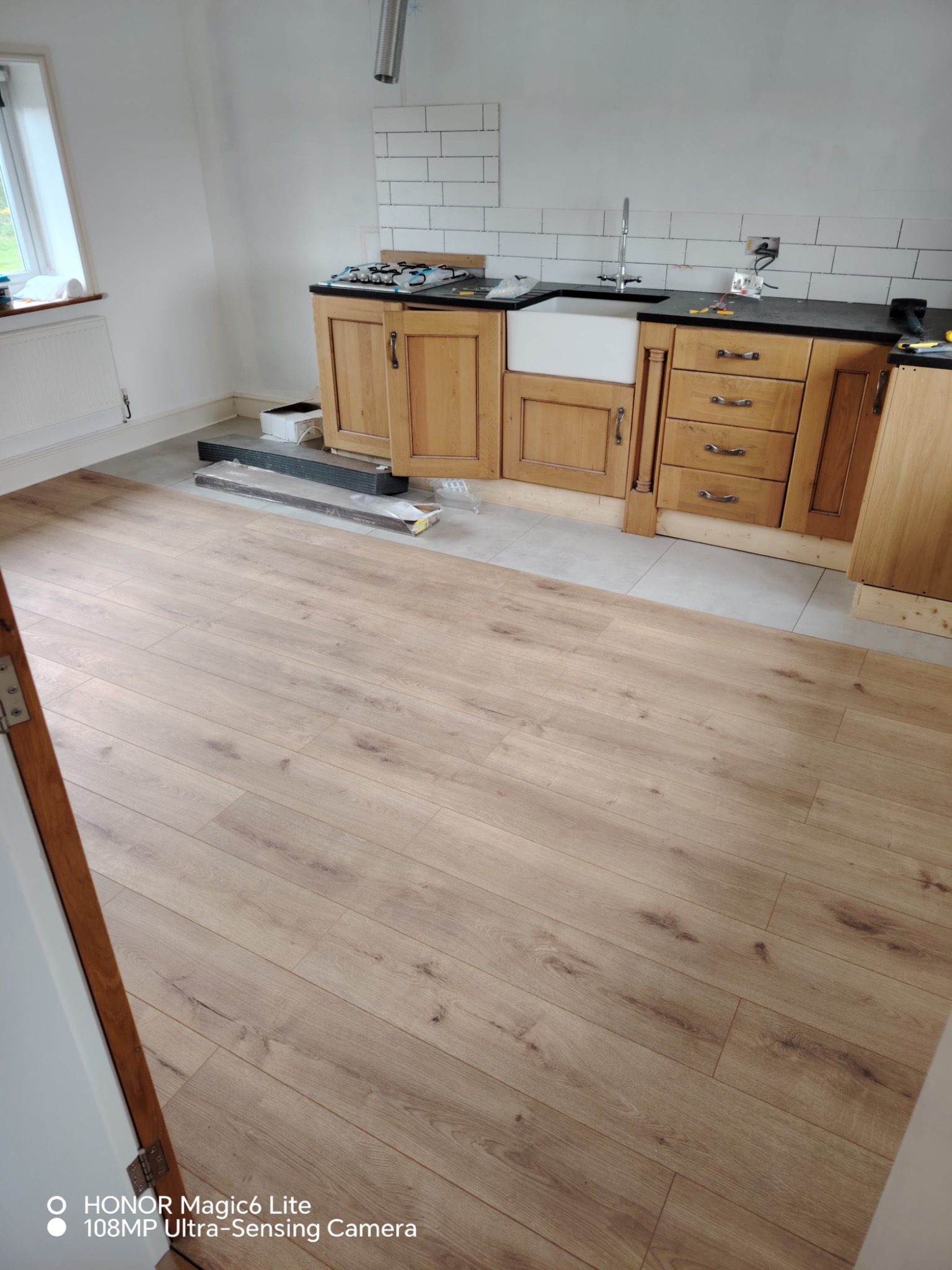 Jasne panele podłogowe w kuchni z drewnianymi szafkami i białymi płytkami na ścianie. Częściowo ułożona podłoga, widoczne paczki paneli i narzędzia.