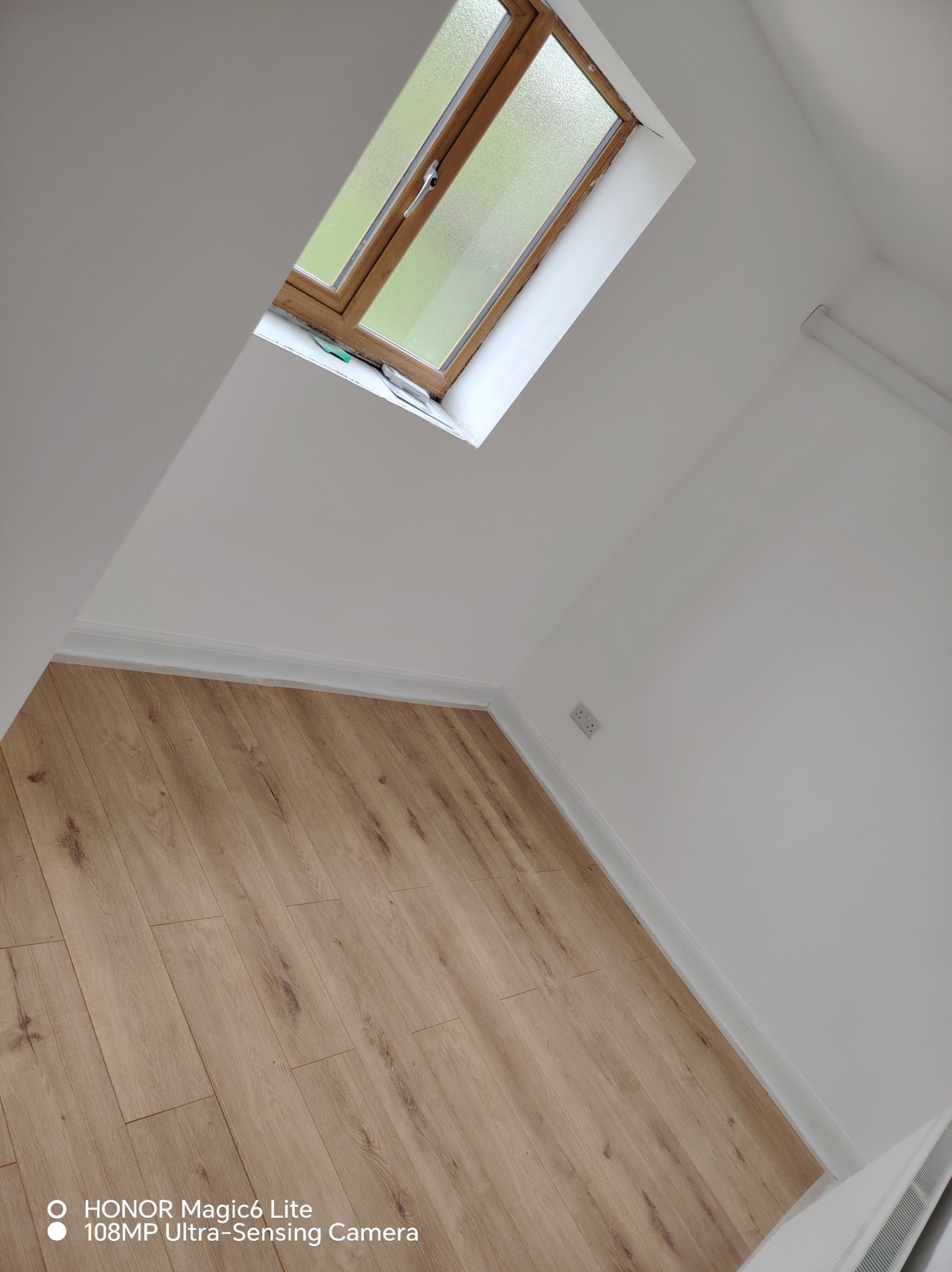 Jasny pokój na poddaszu po remoncie: drewniana podłoga, białe ściany, okno dachowe z widokiem na zieleń. Widoczne gniazdko elektryczne i listwy przypodłogowe.