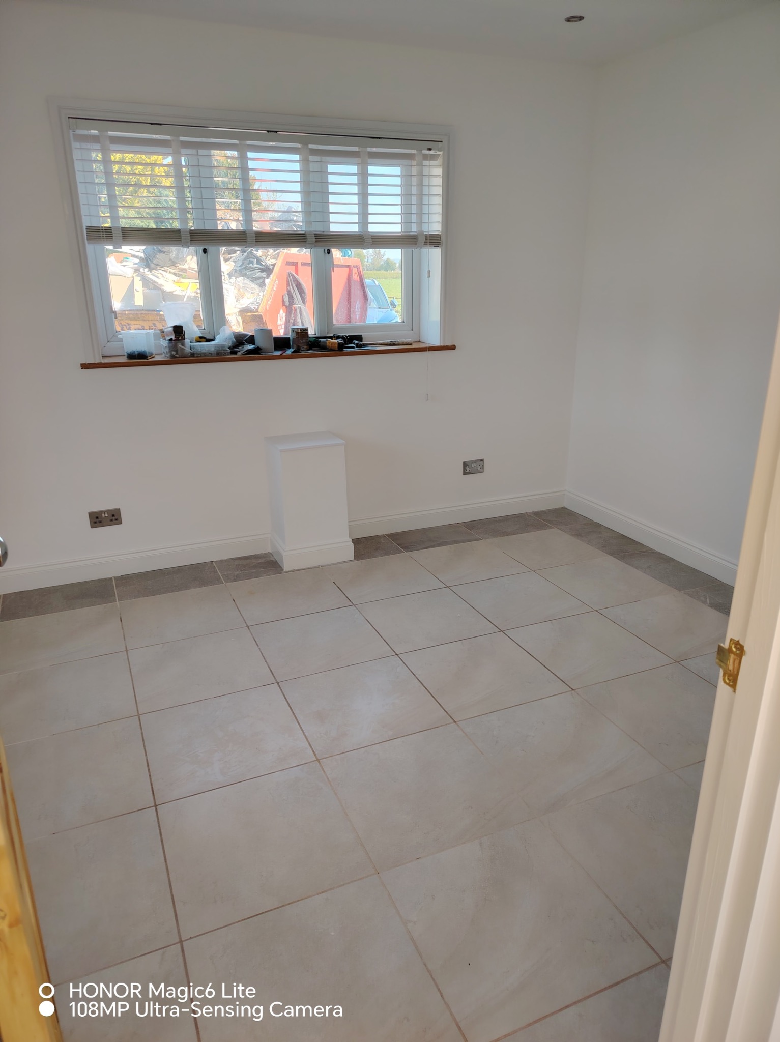 Jasne wnętrze po remoncie: beżowe płytki podłogowe, białe ściany z listwami przypodłogowymi. Okno z żaluzjami, na parapecie narzędzia i akcesoria.