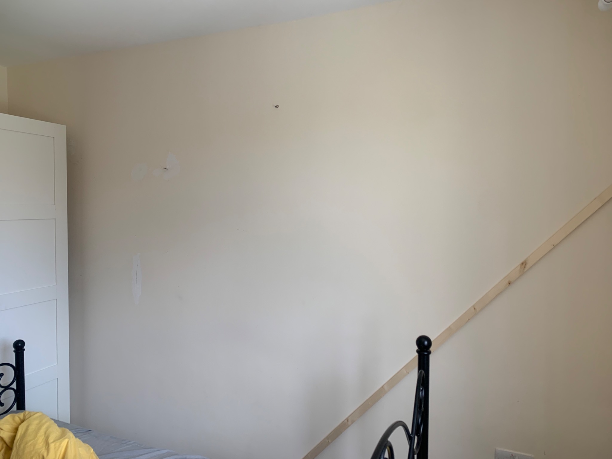 Ściana w pokoju przed malowaniem: widoczne ślady po szpachlowaniu, gdzieniegdzie dziury i listwa przypodłogowa. Przy ścianie fragment łóżka i szafy.