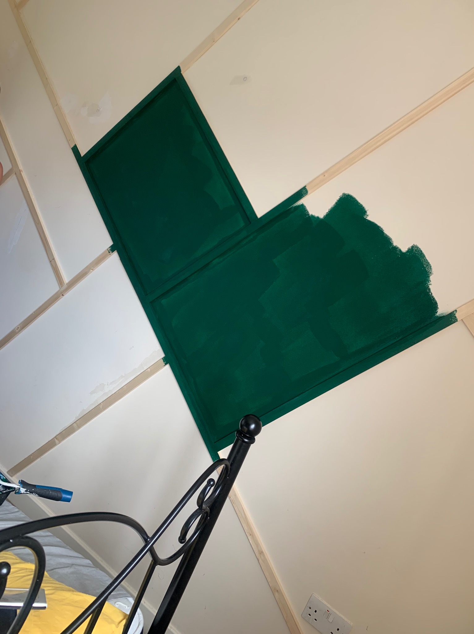 Fragment pokoju w trakcie malowania na zielono, widoczne drewniane listwy i część metalowego łóżka. Ściana pomalowana częściowo, ukazująca etap prac.