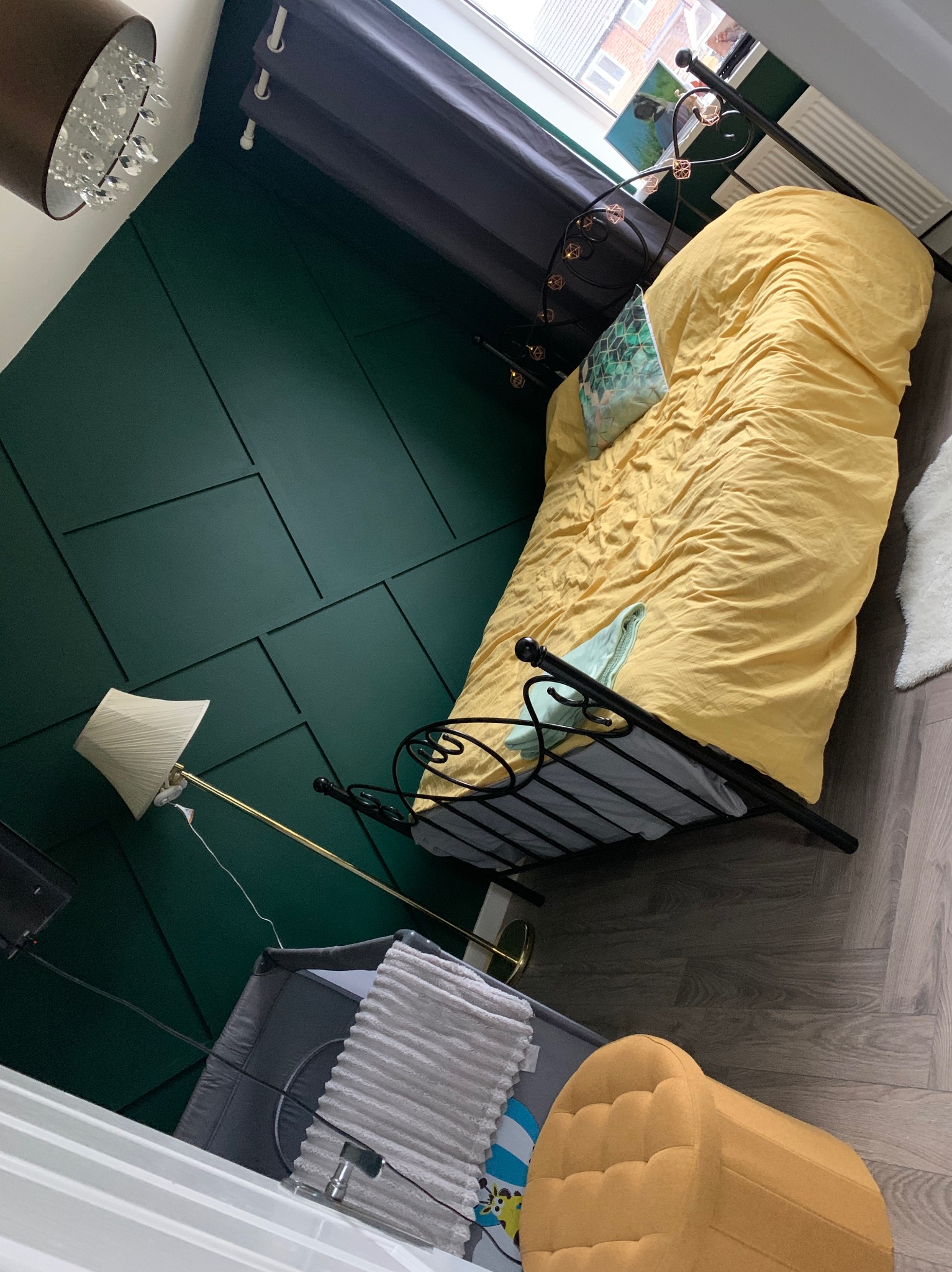 Pokój z łóżkiem, zieloną ścianą z panelami i lampą. Widoczne detale wystroju, takie jak żyrandol i zasłony, tworzą przytulną atmosferę.