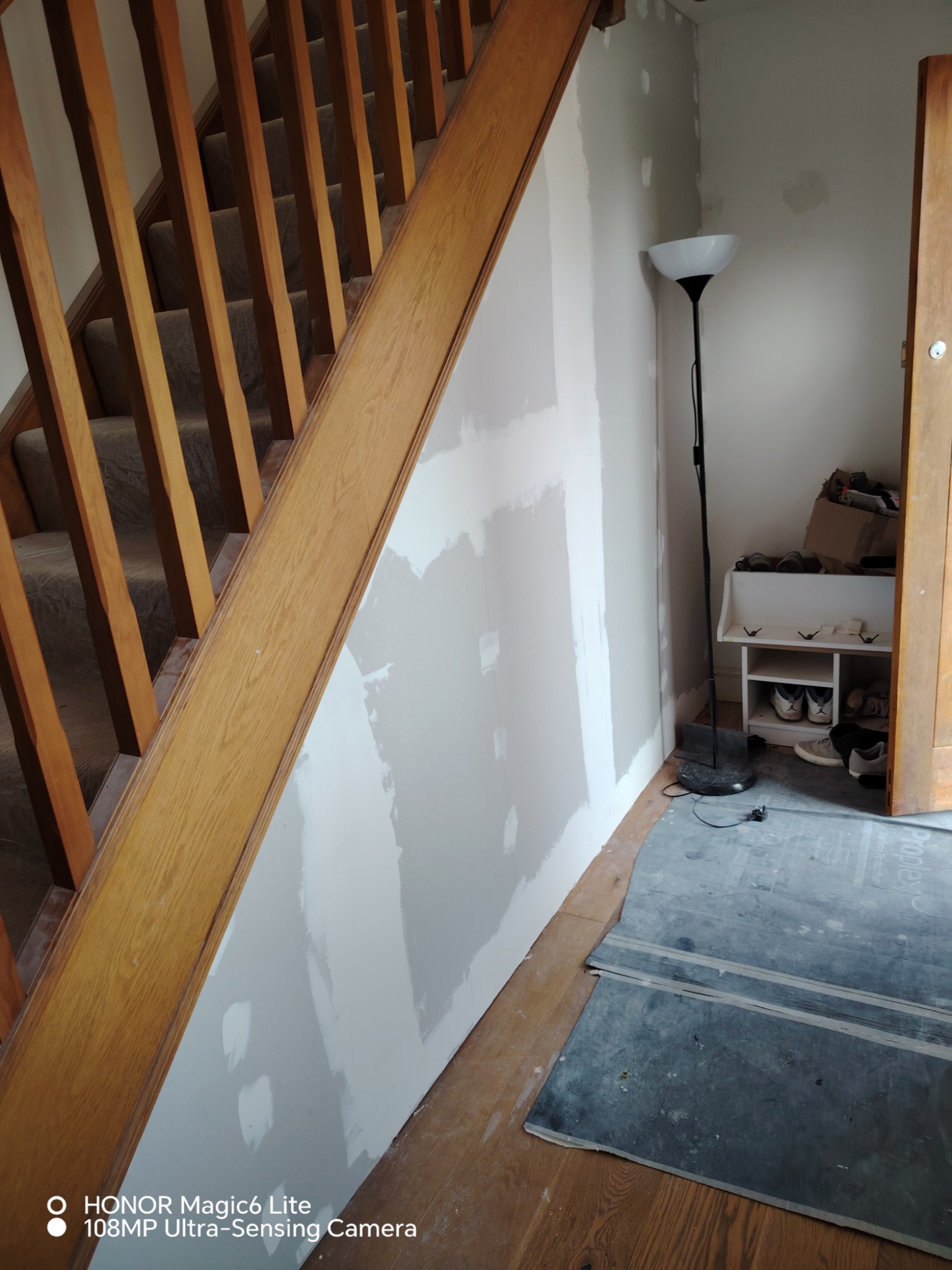 Ściana po szpachlowaniu przy drewnianych schodach, widoczne niedoskonałości. Podłoga zabezpieczona, w tle lampa i szafka na buty. Wnętrze w trakcie remontu.