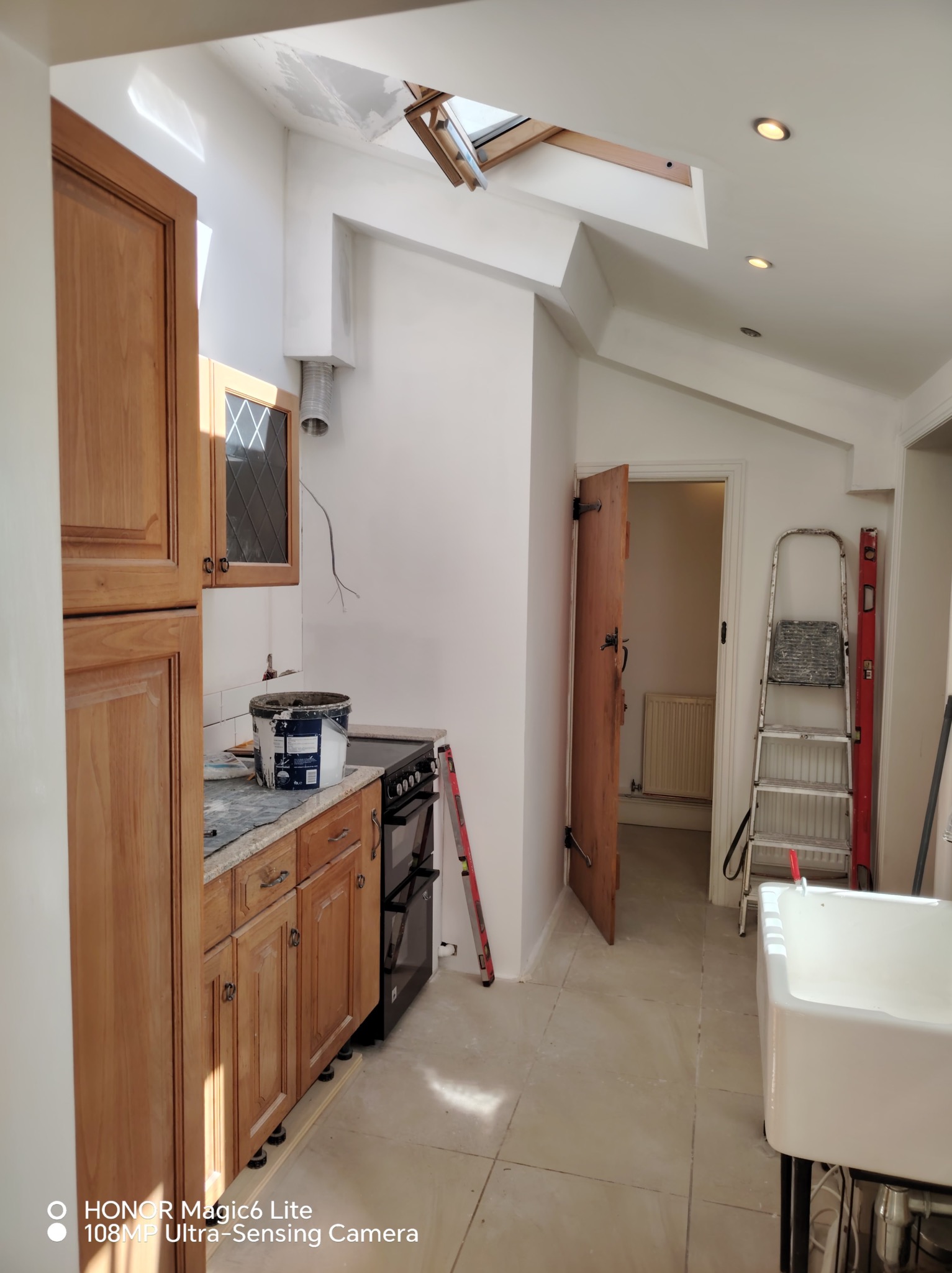 Wnętrze kuchni w trakcie remontu: drewniane szafki, białe ściany, drabina, otwarte okno dachowe. Widoczne ślady prac wykończeniowych i narzędzia.