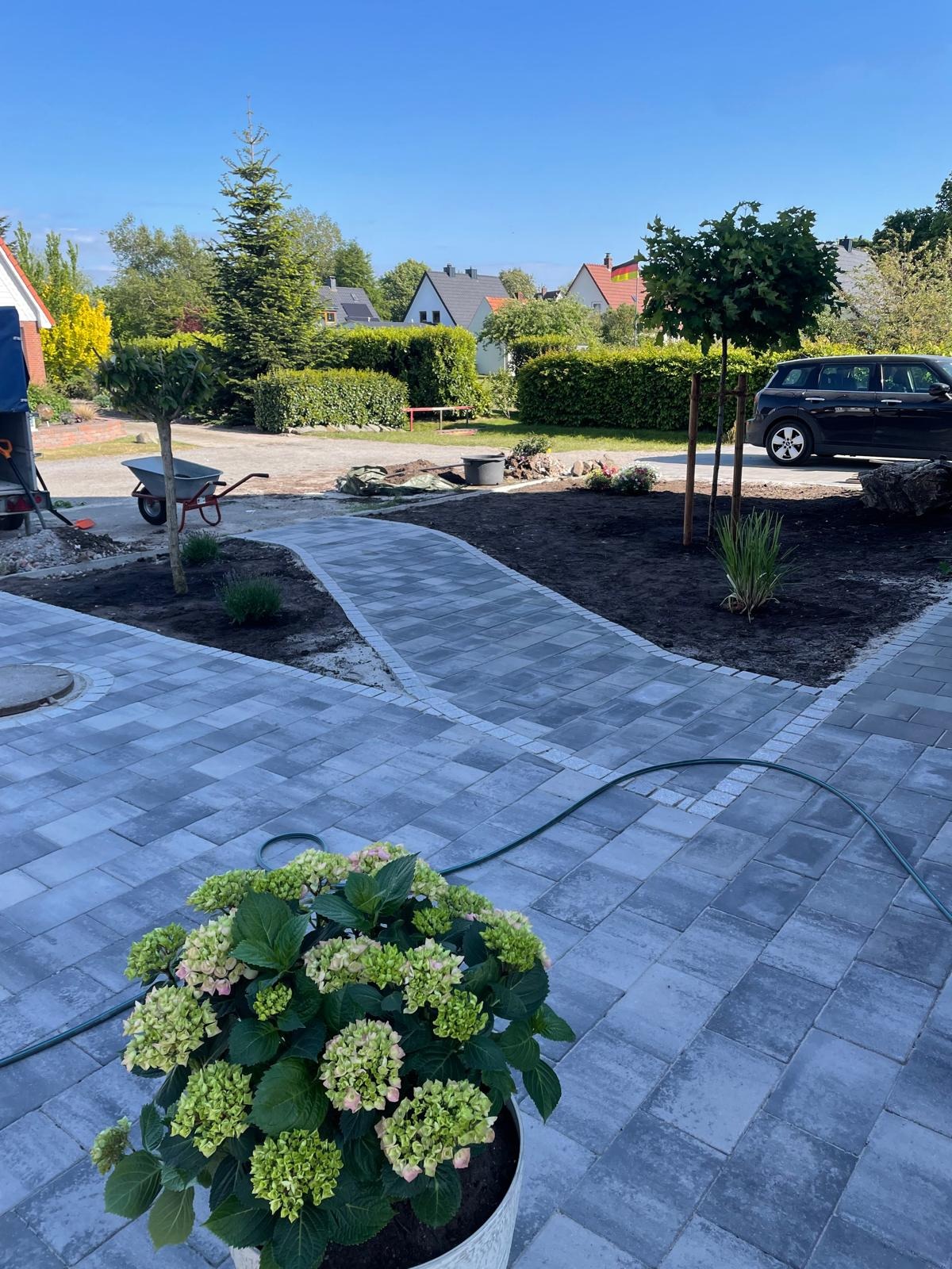 Ścieżka z kostki brukowej w ogrodzie, z obrzeżami i kwiatami w donicy. W tle drzewa, krzewy i domy. Widoczny wózek i narzędzia ogrodnicze.