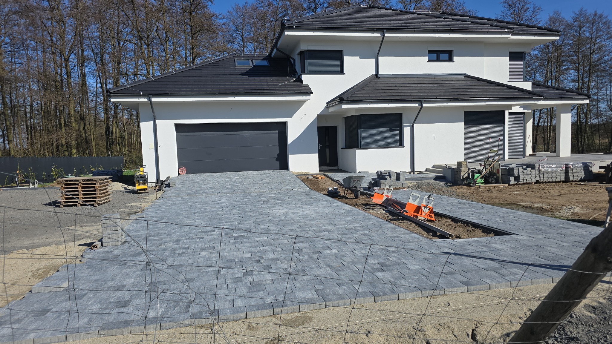 Nowa, szara kostka brukowa ułożona na podjeździe do nowoczesnego domu z ciemną dachówką. Widoczne narzędzia brukarskie i materiały budowlane. Częściowo ukończony projekt.