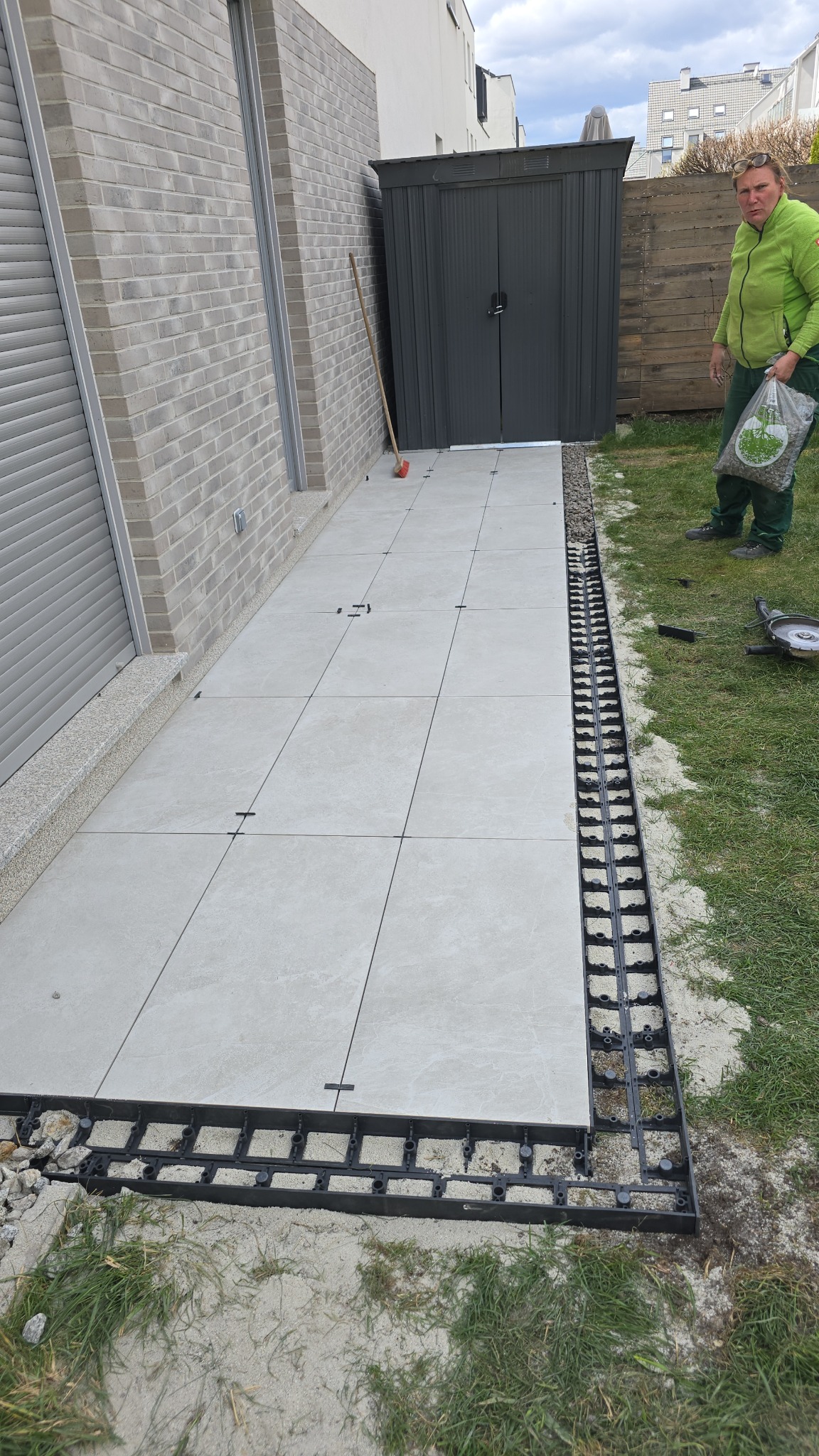 Montaż tarasu z płyt na plastikowych legarach. Widoczny fragment ułożonych płyt, legary i osoba z workiem. W tle budynek z szarymi roletami i szopa.