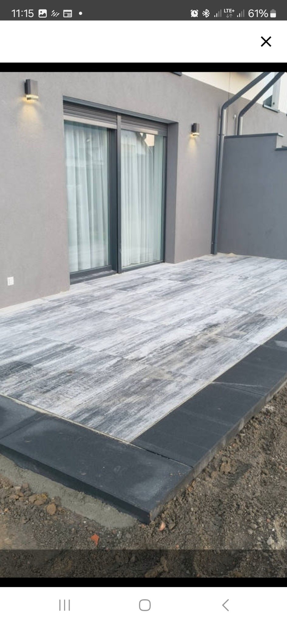 Ułożona kostka brukowa w odcieniach szarości na tarasie przydomowym, z ciemnoszarą obwódką. Widoczne oświetlenie elewacyjne i fragment schodów.