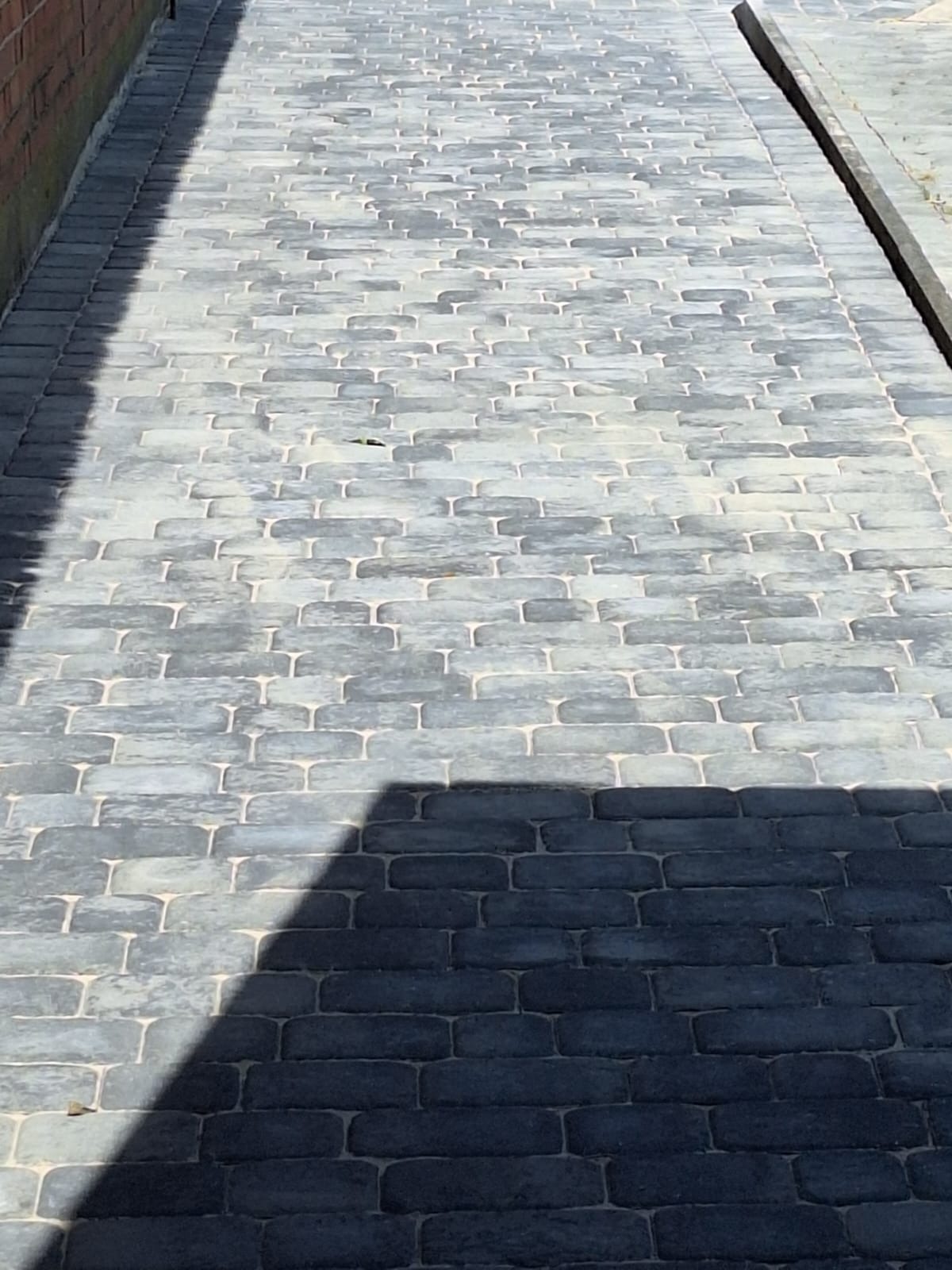 Szara kostka brukowa ułożona w regularny wzór, oświetlona częściowo słońcem, tworząc cień na fragmencie powierzchni. Widoczna krawędź chodnika i ściana.