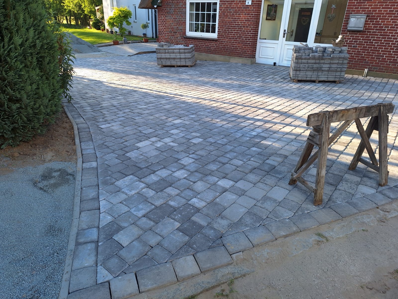 Ułożona kostka brukowa w odcieniach szarości tworząca wzór na podjeździe. Paleta z kostką i drewniany kozioł na pierwszym planie. Widok na dom z czerwonej cegły.