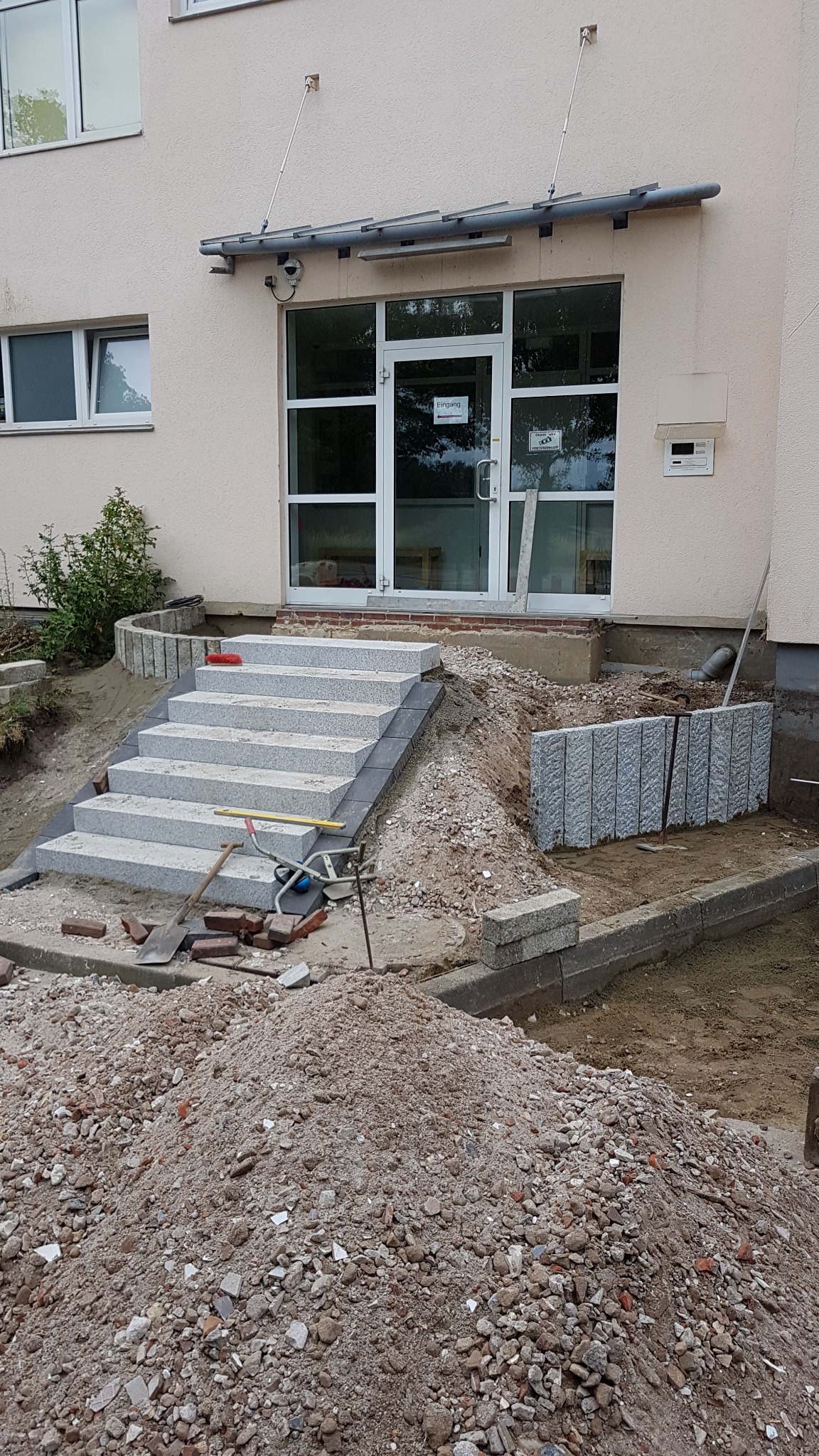 Montaż schodów z kostki granitowej przed budynkiem. Widoczne narzędzia, piasek i częściowo ułożone obrzeża. Prace w toku.