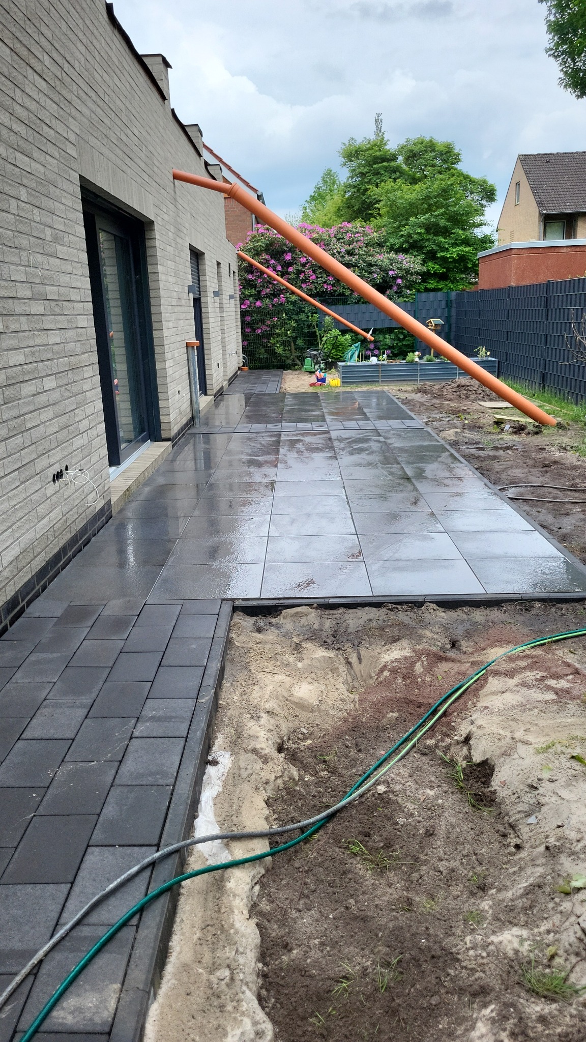 Częściowo ułożona, mokra, szara kostka brukowa na tarasie, z widocznym przygotowanym podłożem. Rury spustowe i zielony wąż ogrodowy w kadrze.