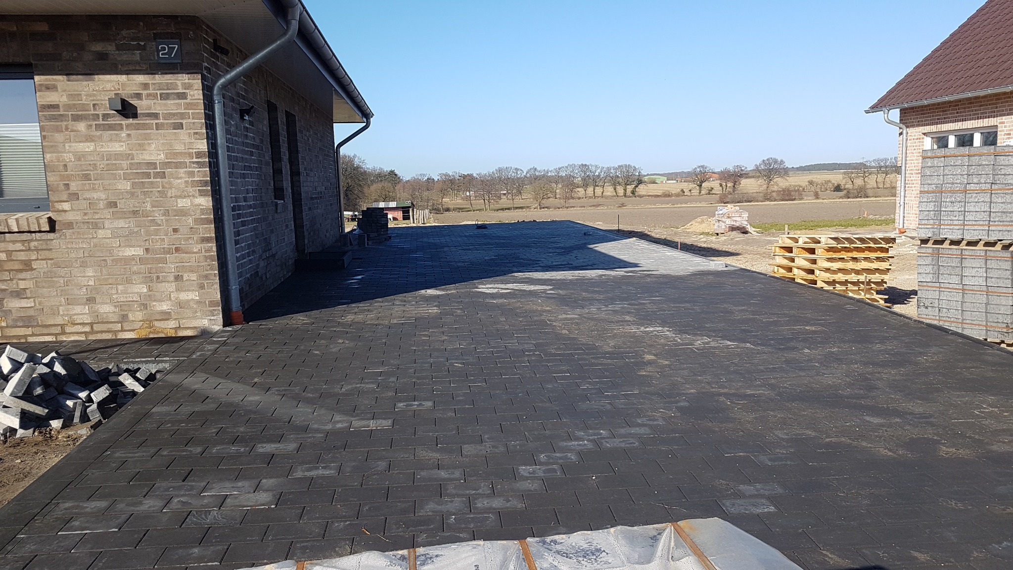 Ciemnoszara kostka brukowa ułożona przed budynkiem z cegły, palety z kostką w tle, widok na pole. Częściowo ułożona nawierzchnia z kostki.