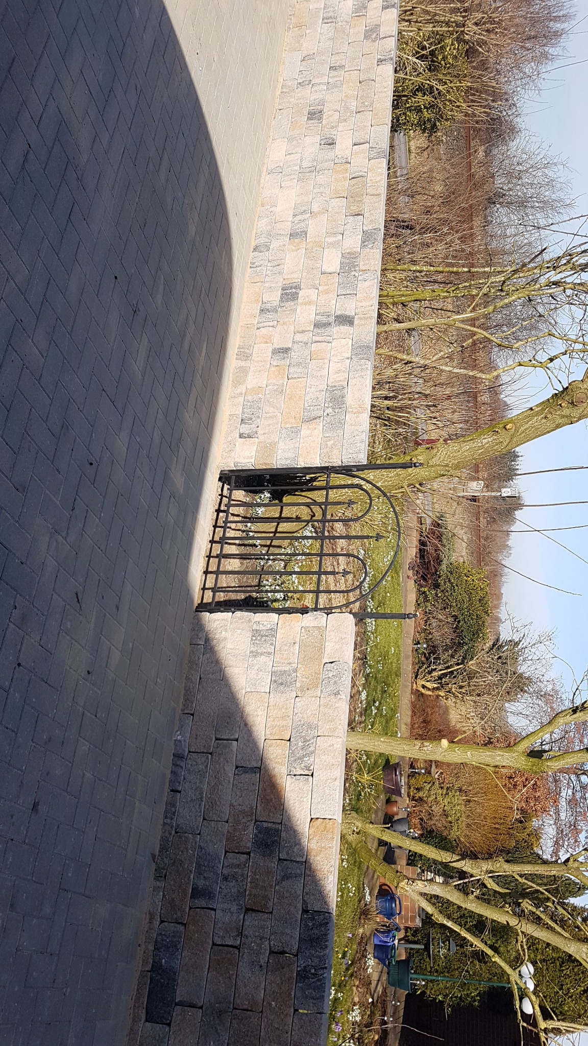 Brukowa ścieżka z dekoracyjną, niską, metalową bramką ogrodową, obok murku z kamienia. W tle zieleń i drzewa wczesną wiosną.