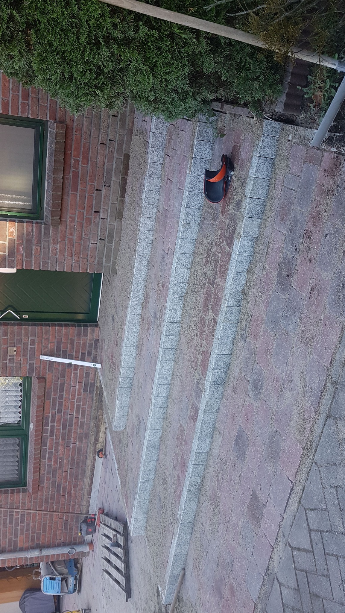 Schody z kostki brukowej z szarymi obrzeżami prowadzące do domu z cegły, widoczne narzędzia i poziomica. Układanie kostki brukowej w toku.