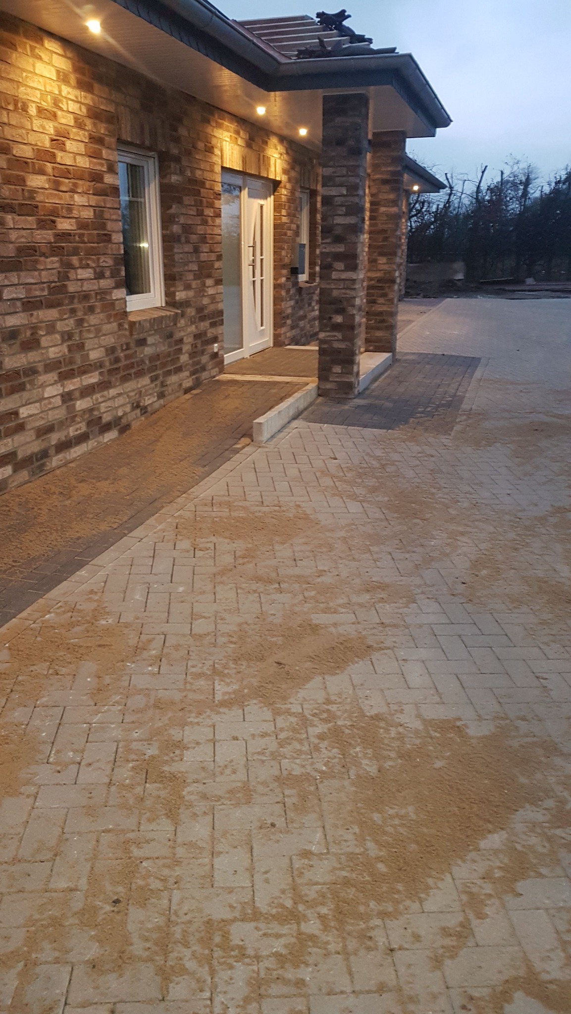 Ułożona kostka brukowa przed domem z cegły klinkierowej, oświetlona lampami. Widoczny podjazd z kostki i fragment ogrodu. Całość w stonowanej kolorystyce.