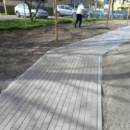 Goldenbruk - Świeżo ułożona, szara kostka brukowa tworzy ścieżkę wzdłuż trawnika. W tle widoczna osoba podczas prac wykończeniowych i zaparkowane samochody.