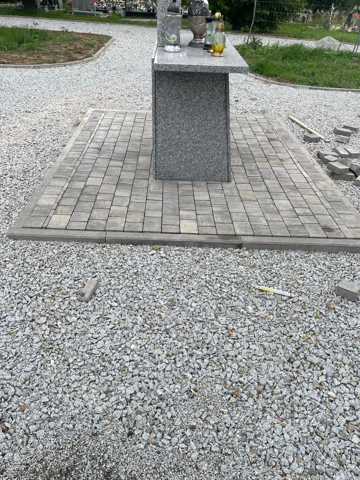 Szara kostka brukowa ułożona wokół pomnika na cmentarzu, z rozrzuconymi kostkami i żwirem. W tle nagrobki i zieleń. Widok z góry.