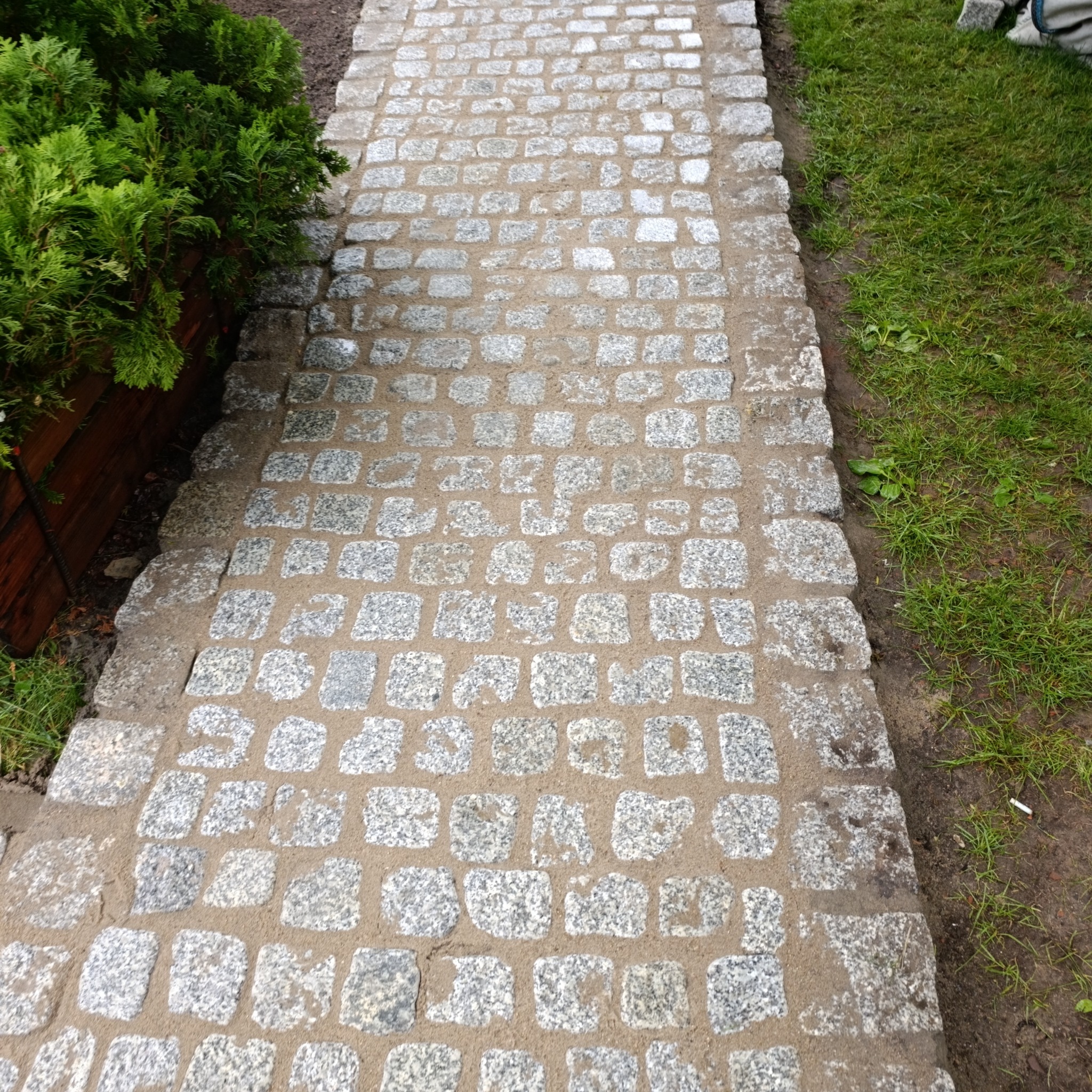 Ścieżka z jasnoszarej kostki granitowej, wypełniona piaskiem, obok trawnik i krzewy. Widok z góry, na ukośnej osi.