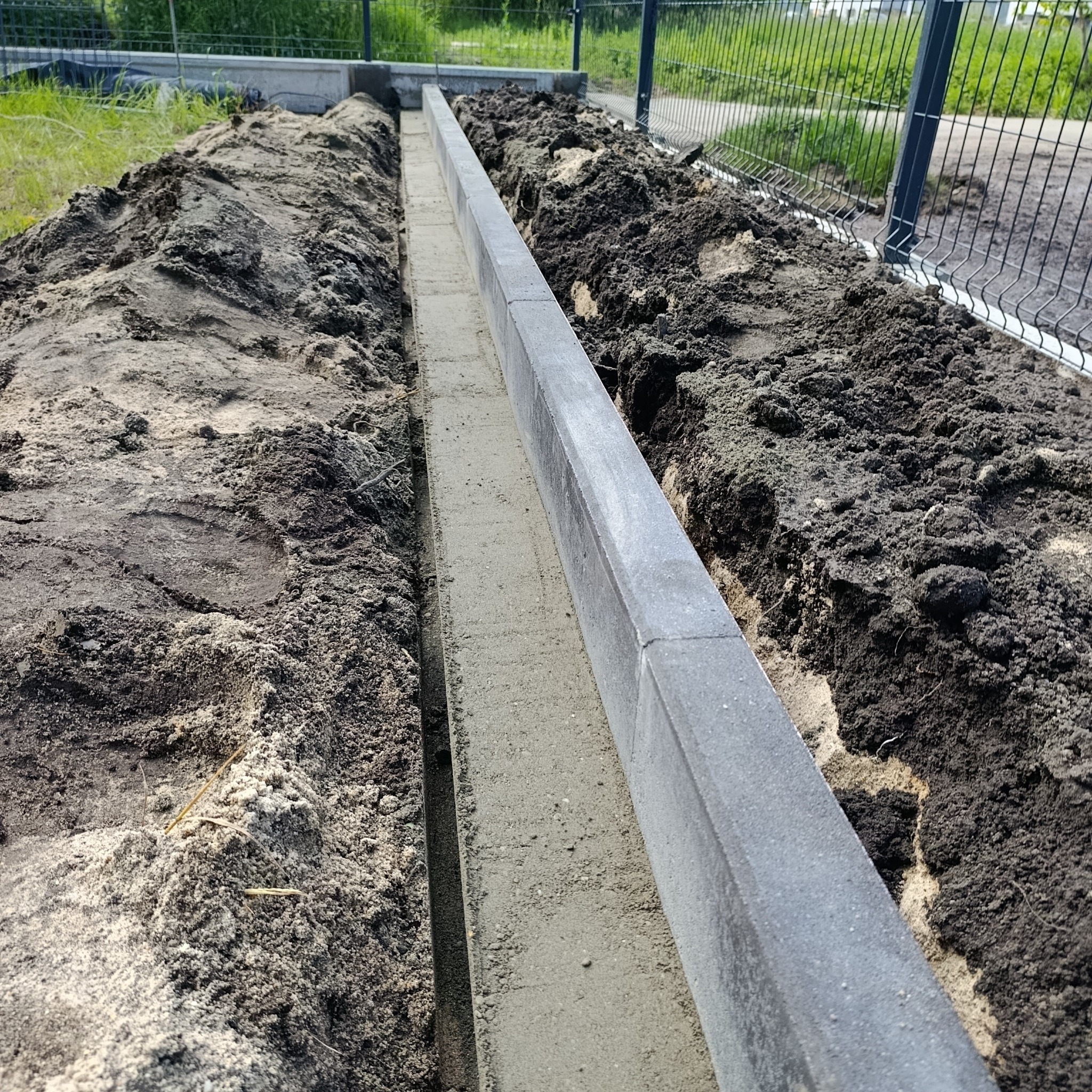 Wykop z betonową wylewką i obrzeżami z kostki, ziemia po bokach, siatka ogrodzeniowa w tle. Przygotowanie podłoża pod ścieżkę lub podjazd.