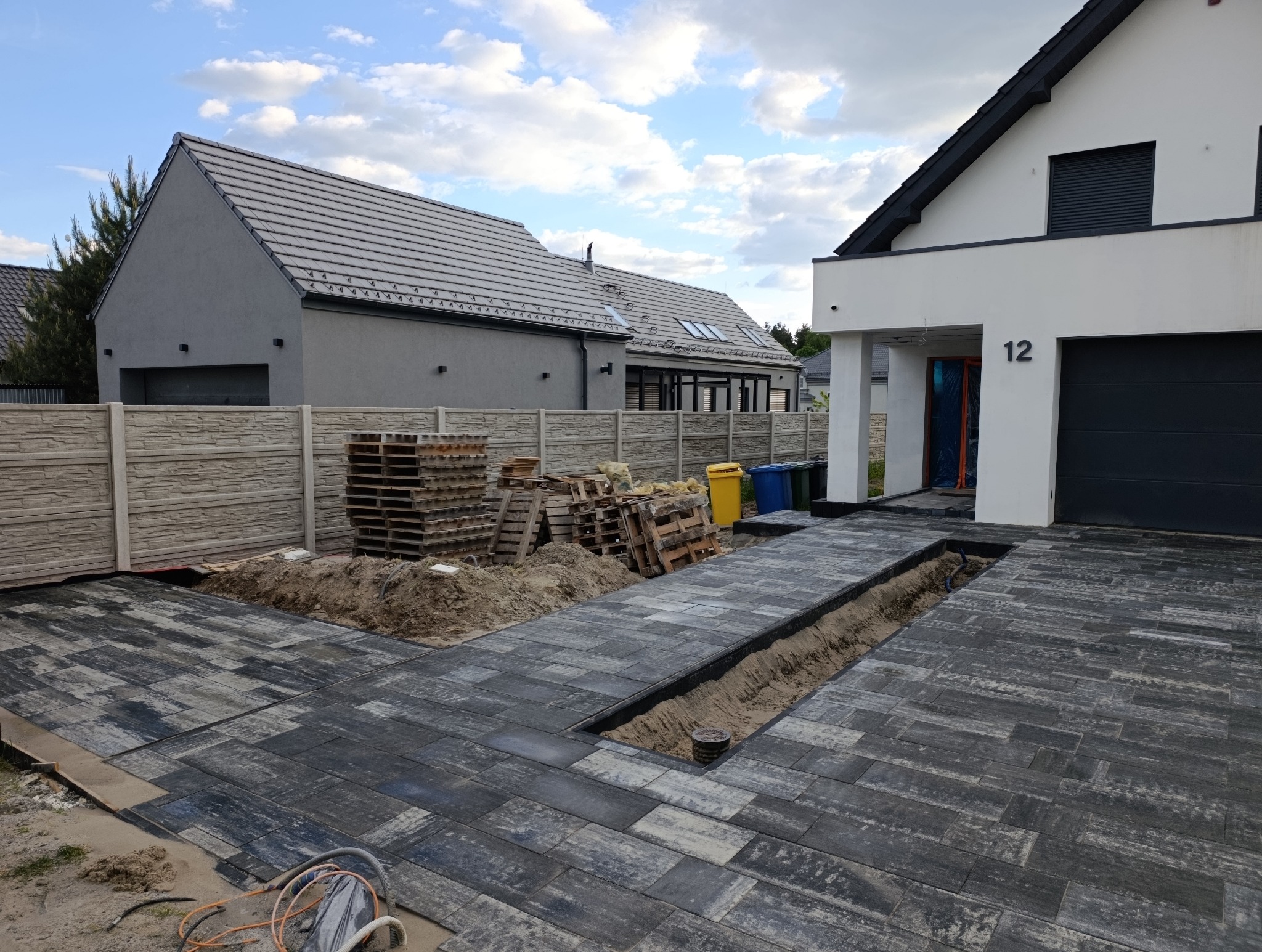 Częściowo ułożona, szara kostka brukowa na podjeździe domu z numerem 12, widoczne palety z materiałem i wykop pod instalacje. Słoneczny dzień.