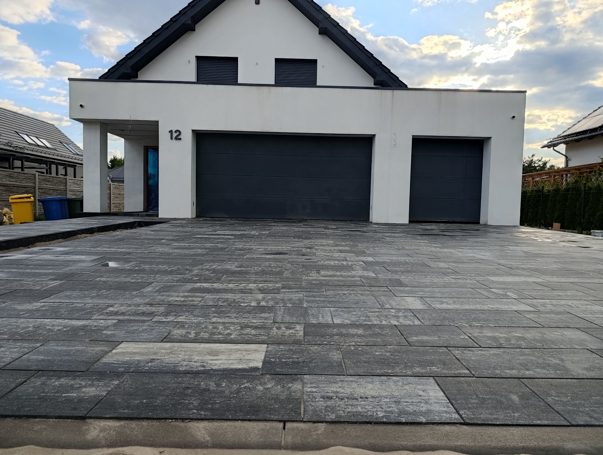 Nowoczesna posesja z wybrukowanym podjazdem z szarej kostki, widok na dom z dwoma garażami i numerem 12. Kostka ułożona w regularny wzór, w tle niebo z chmurami.