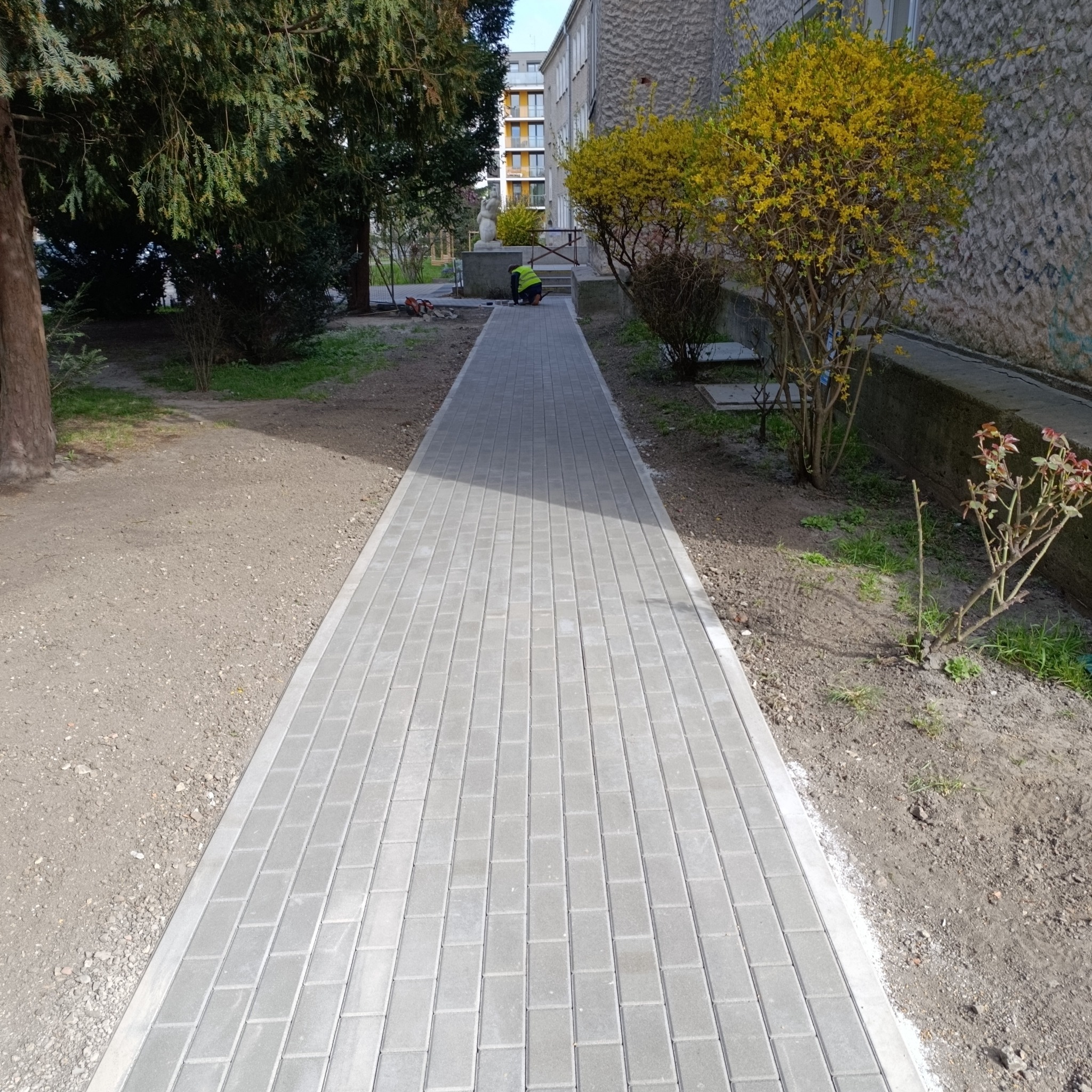 Długa, prosta ścieżka z szarej kostki brukowej, prowadząca wzdłuż budynku. Pracownik w odblaskowej kamizelce w oddali, drzewa i krzewy po bokach.