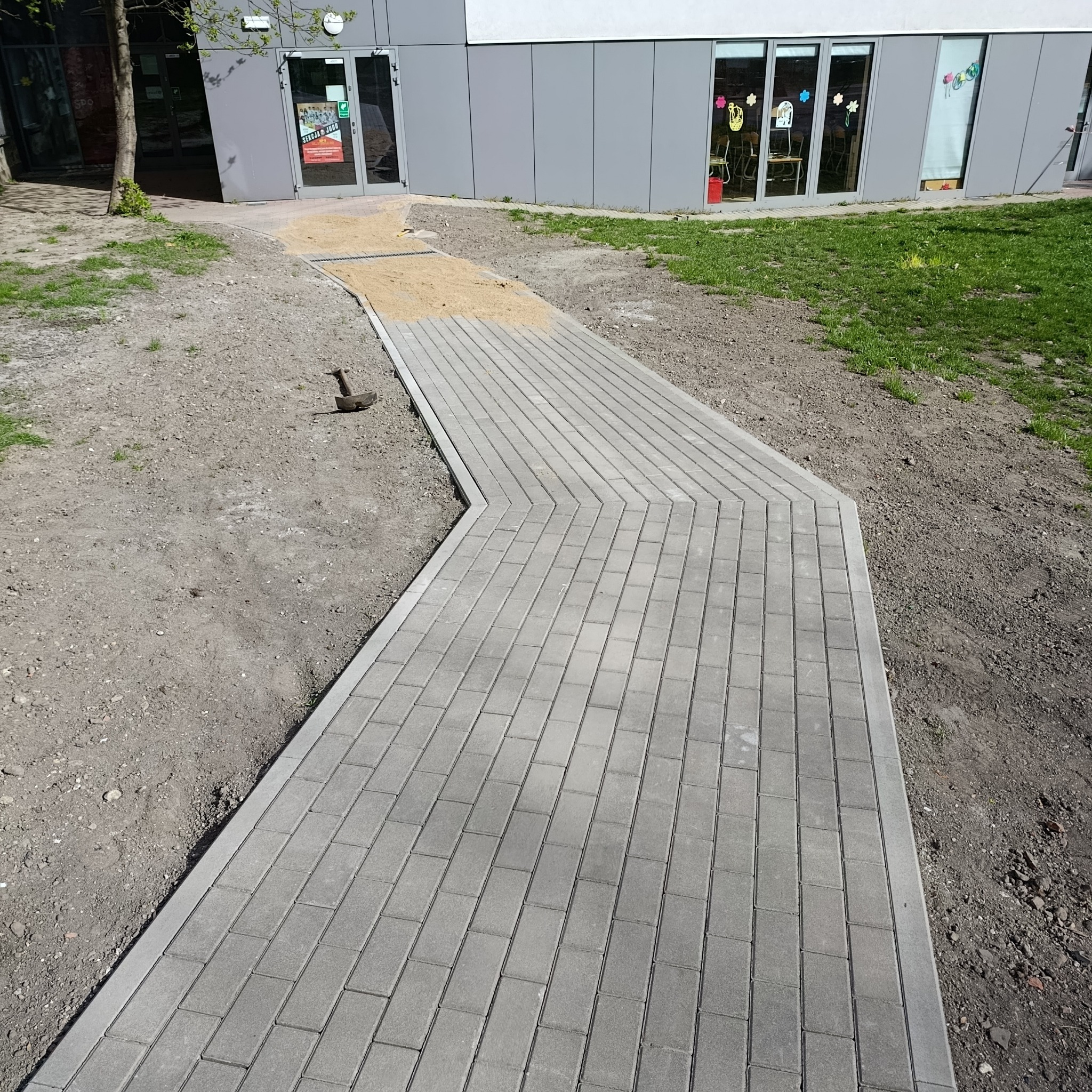 Ścieżka z szarej kostki brukowej prowadząca do budynku, z fragmentem podsypki piaskowej i młotkiem brukarskim obok. Widok z góry.