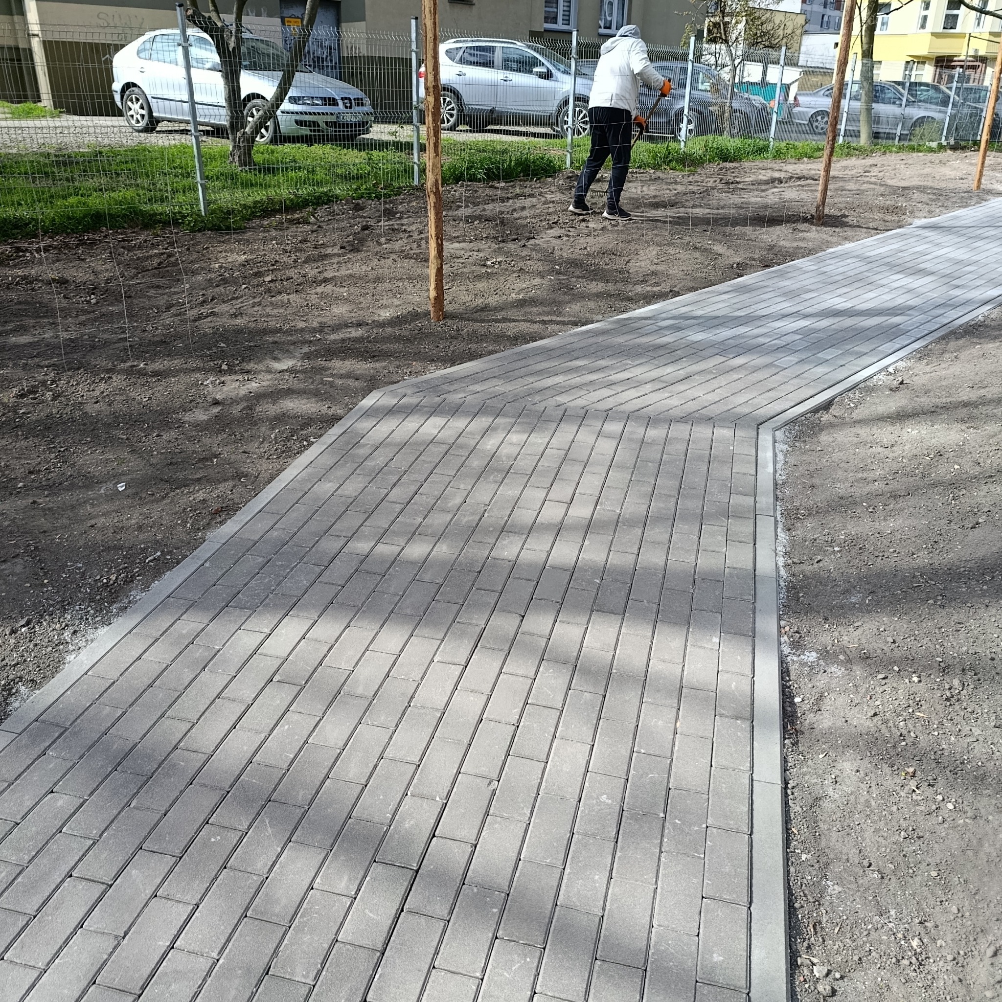 Świeżo ułożona, szara kostka brukowa tworzy ścieżkę wzdłuż trawnika. W tle widoczna osoba podczas prac wykończeniowych i zaparkowane samochody.