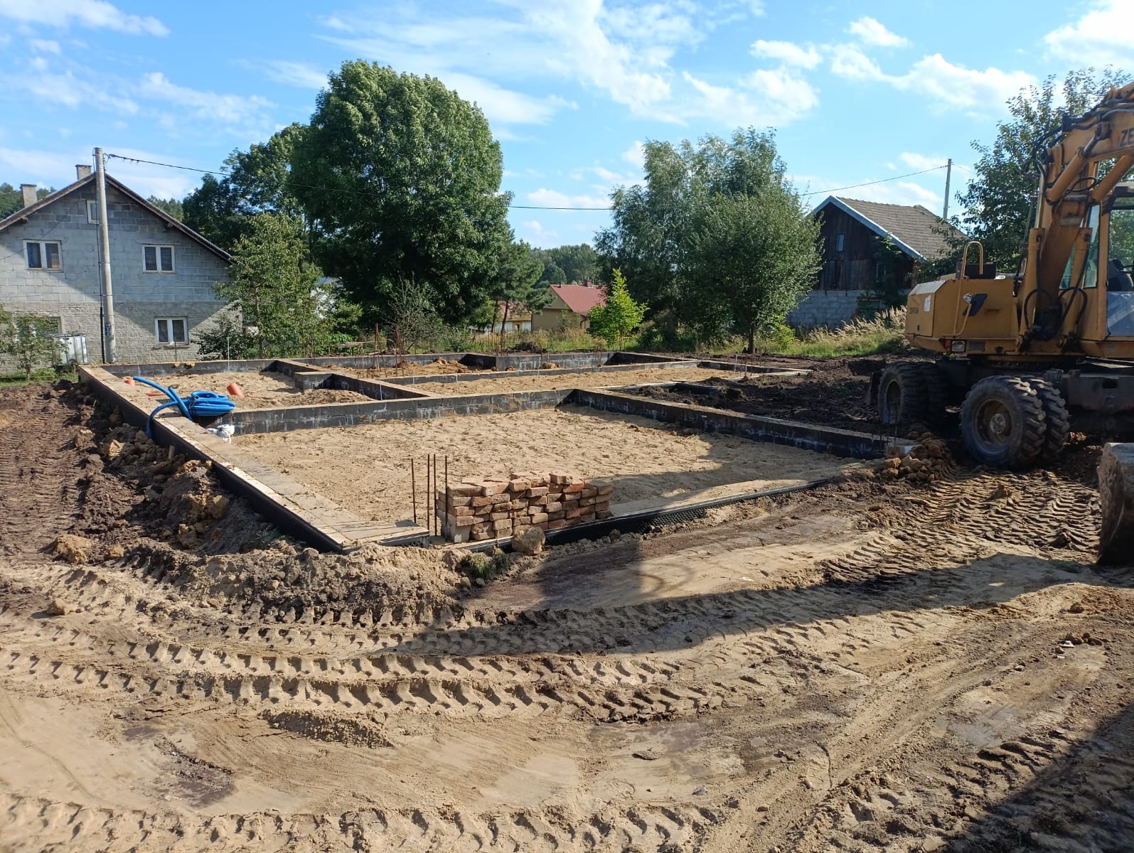 Zalane fundamenty pod budynek jednorodzinny, widoczne szalunki i piasek. Na pierwszym planie ślady opon koparki, w tle dom i drzewa.