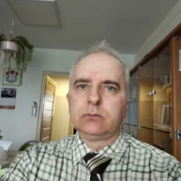 Portret mężczyzny w kraciastej koszuli i krawacie w pomieszczeniu biurowym z szafkami i rośliną doniczkową w tle. Ujęcie z bliska, skupione na twarzy.