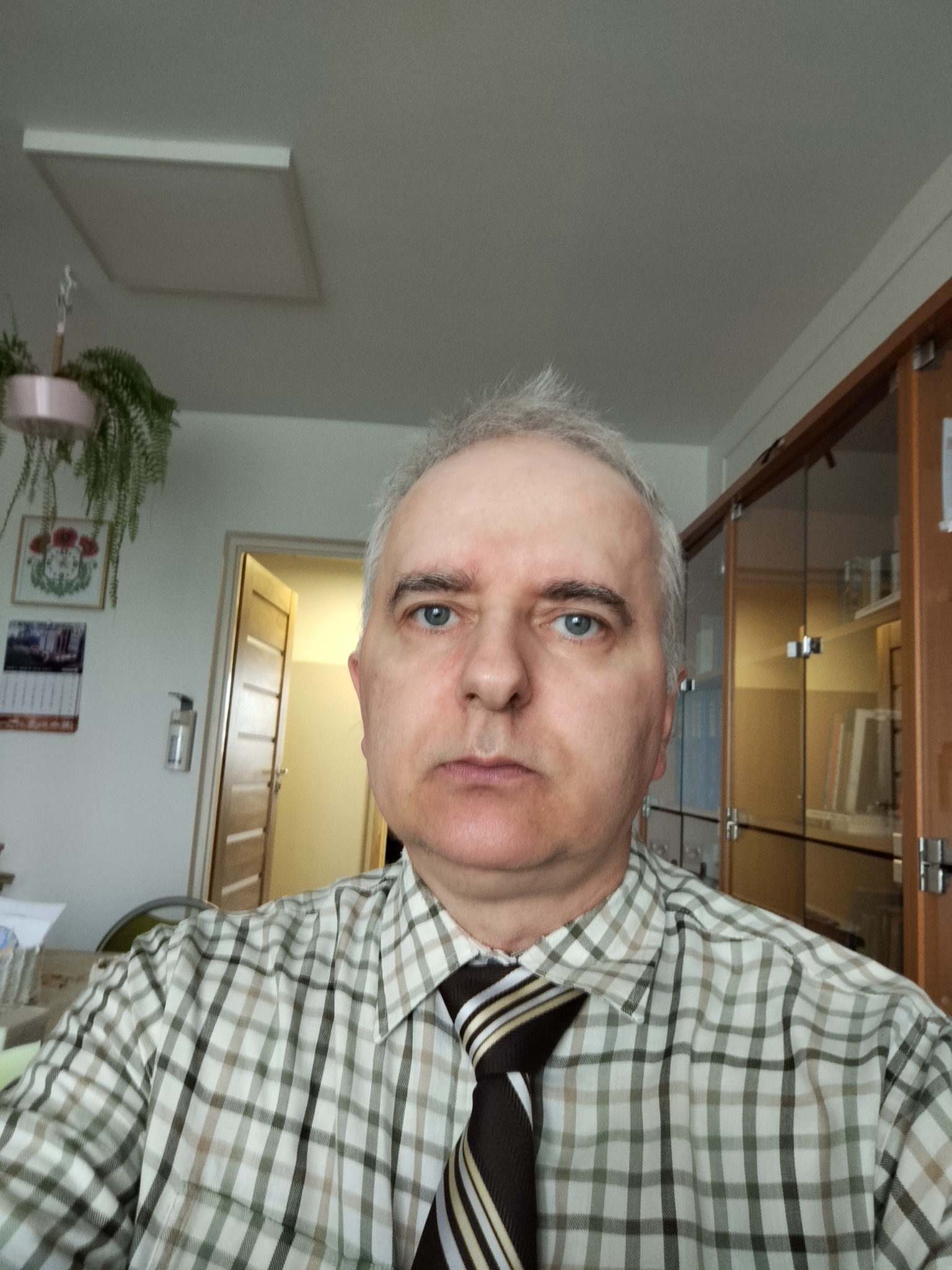 Portret mężczyzny w kraciastej koszuli i krawacie w pomieszczeniu biurowym z szafkami i rośliną doniczkową w tle. Ujęcie z bliska, skupione na twarzy.