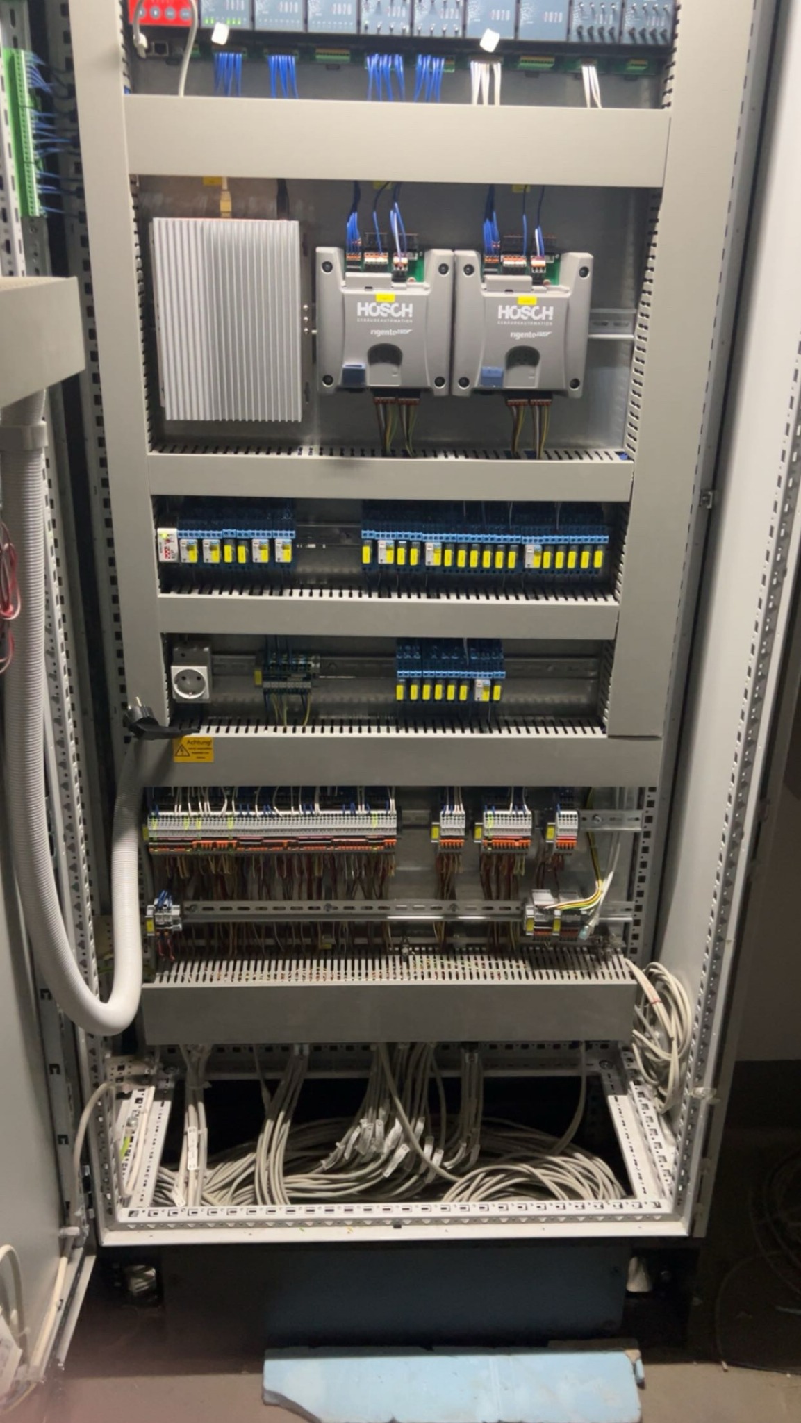 Rozdzielnica elektryczna z licznymi przewodami, bezpiecznikami i elementami sterującymi. Widoczne kable, złączki i urządzenia marki HOSCH.