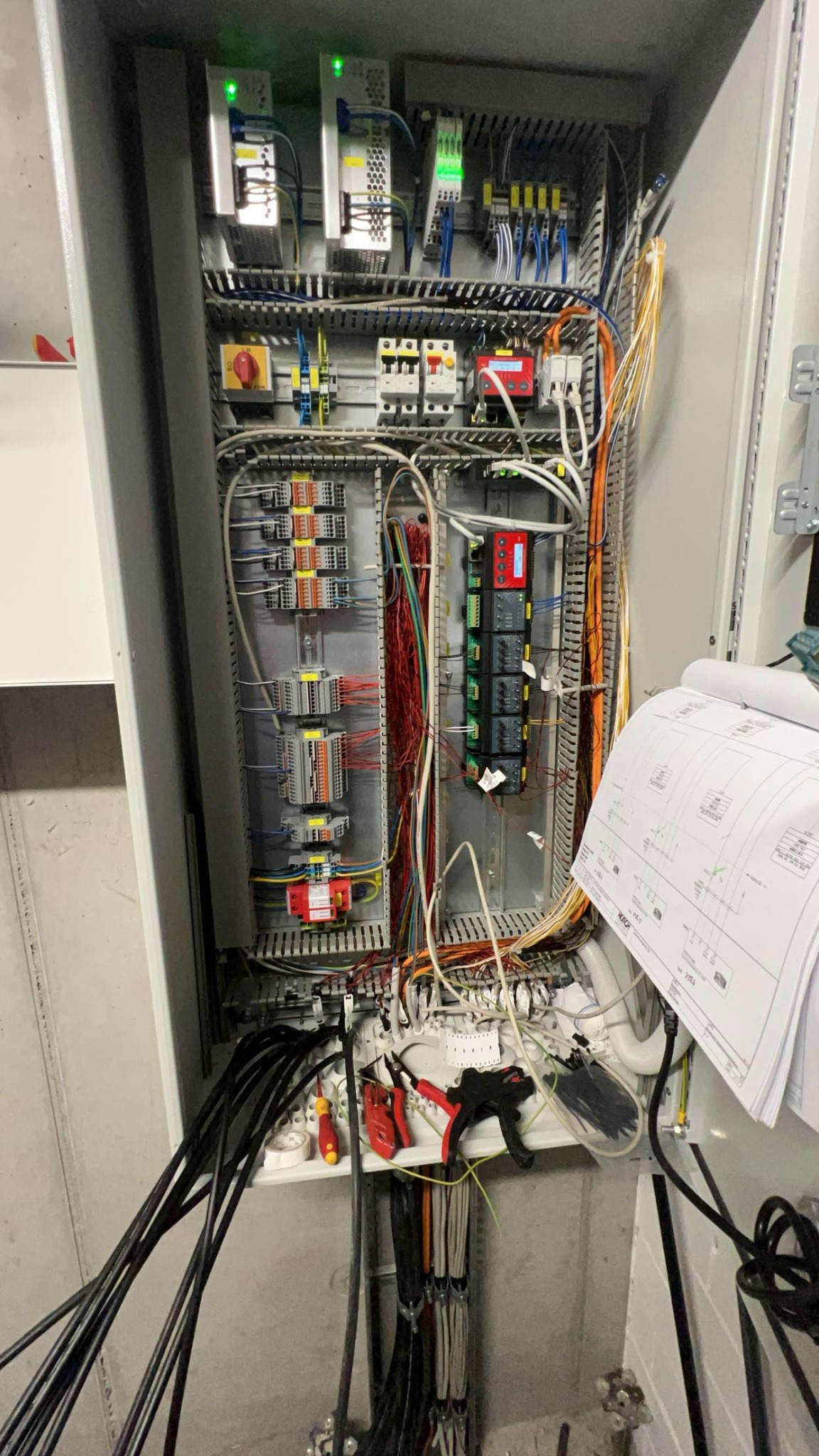 Otwarta szafa sterownicza z widocznymi elementami instalacji elektrycznej, przewodami i narzędziami. Widoczne schematy elektryczne.